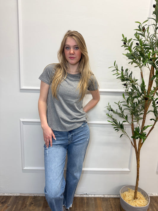 Person wearing a gray t-shirt and blue jeans standing in front of a white door with a plant in the background.