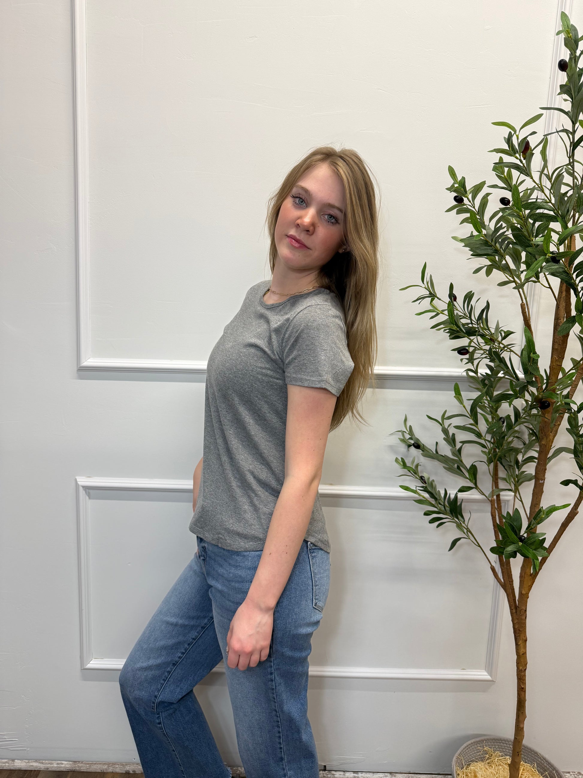 Person wearing a gray shirt and blue jeans standing in front of a white door with a plant in the background.