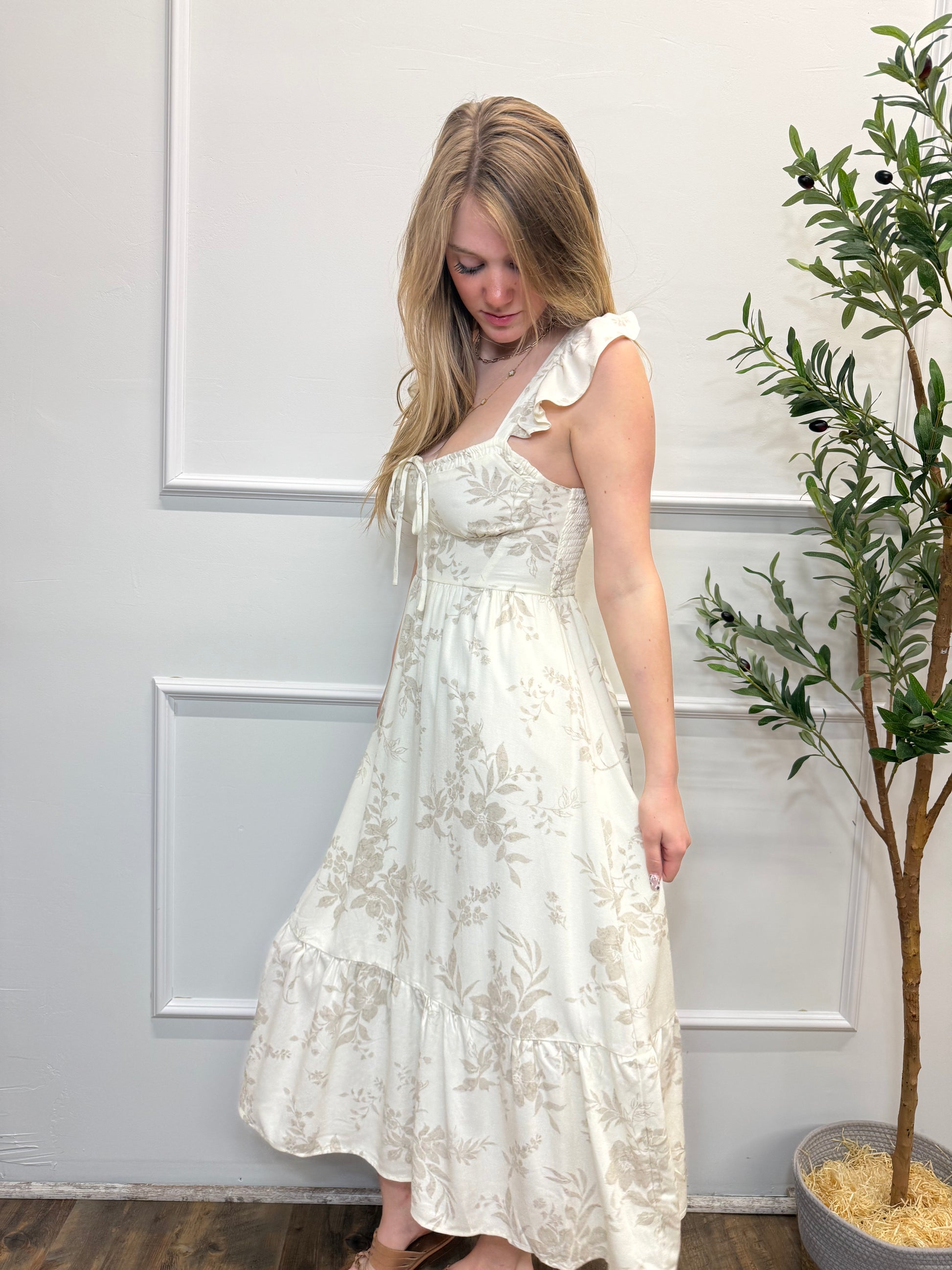 Woman wearing a white lace dress standing against a white wall with a plant in the background.