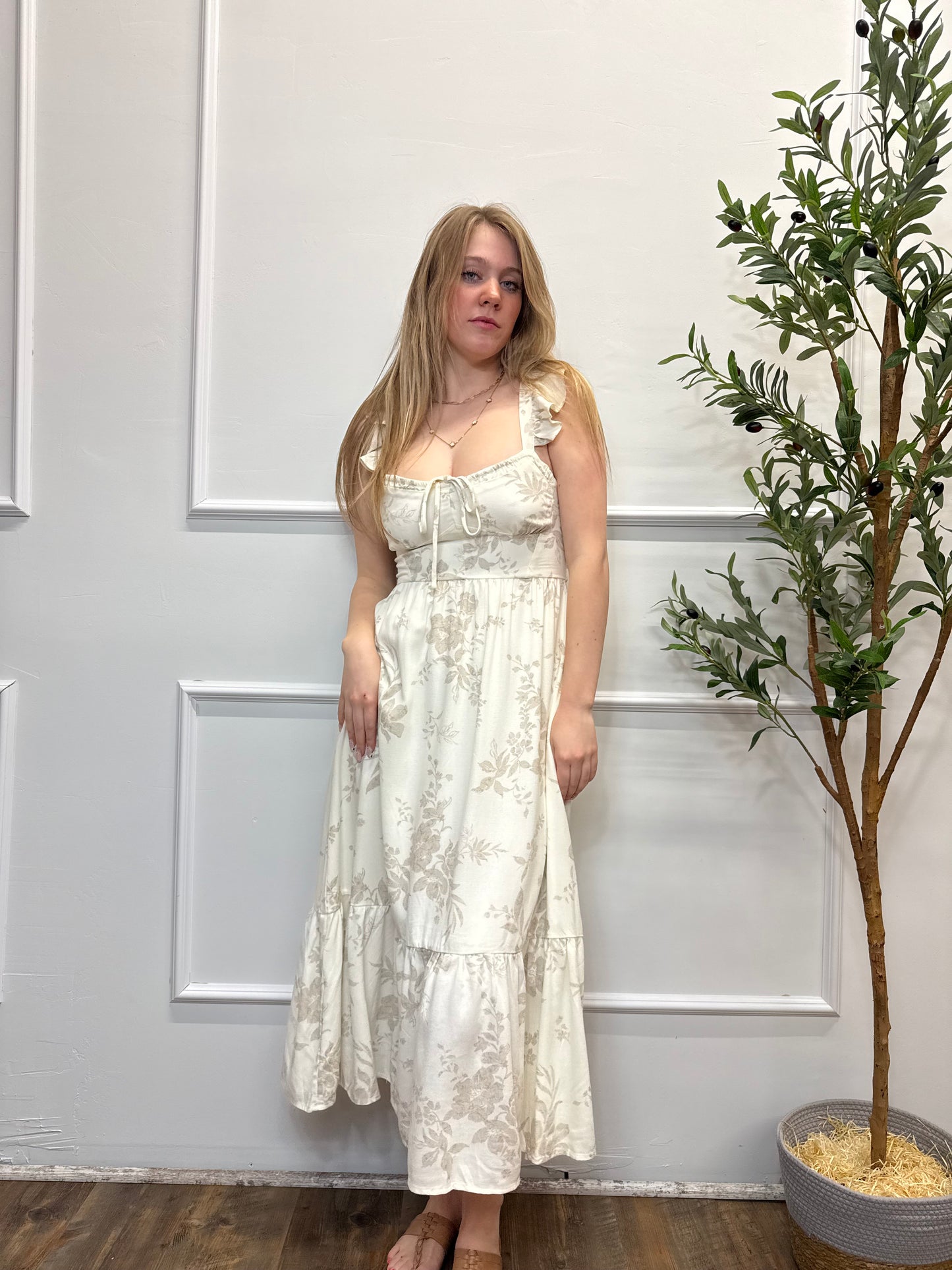 Woman in a white floral dress standing in front of a white door with a plant in the background.