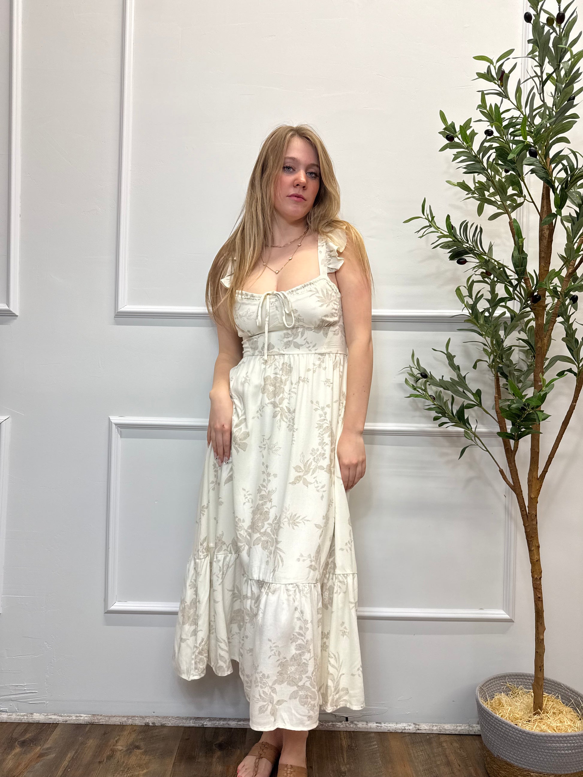 Woman in a white floral dress standing in front of a white door with a plant in the background.