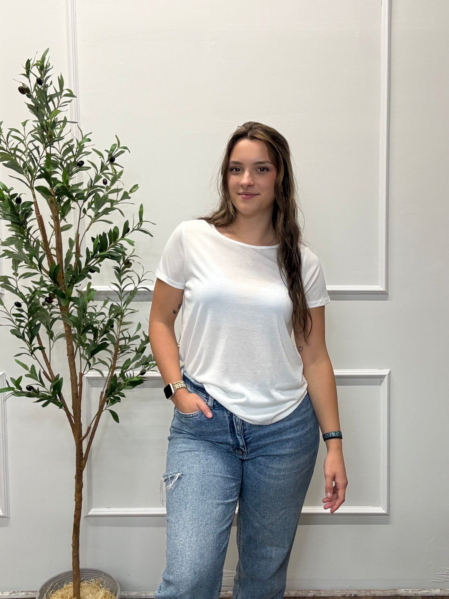 Person wearing a white t-shirt and blue jeans standing in front of a white door with a plant in the background.