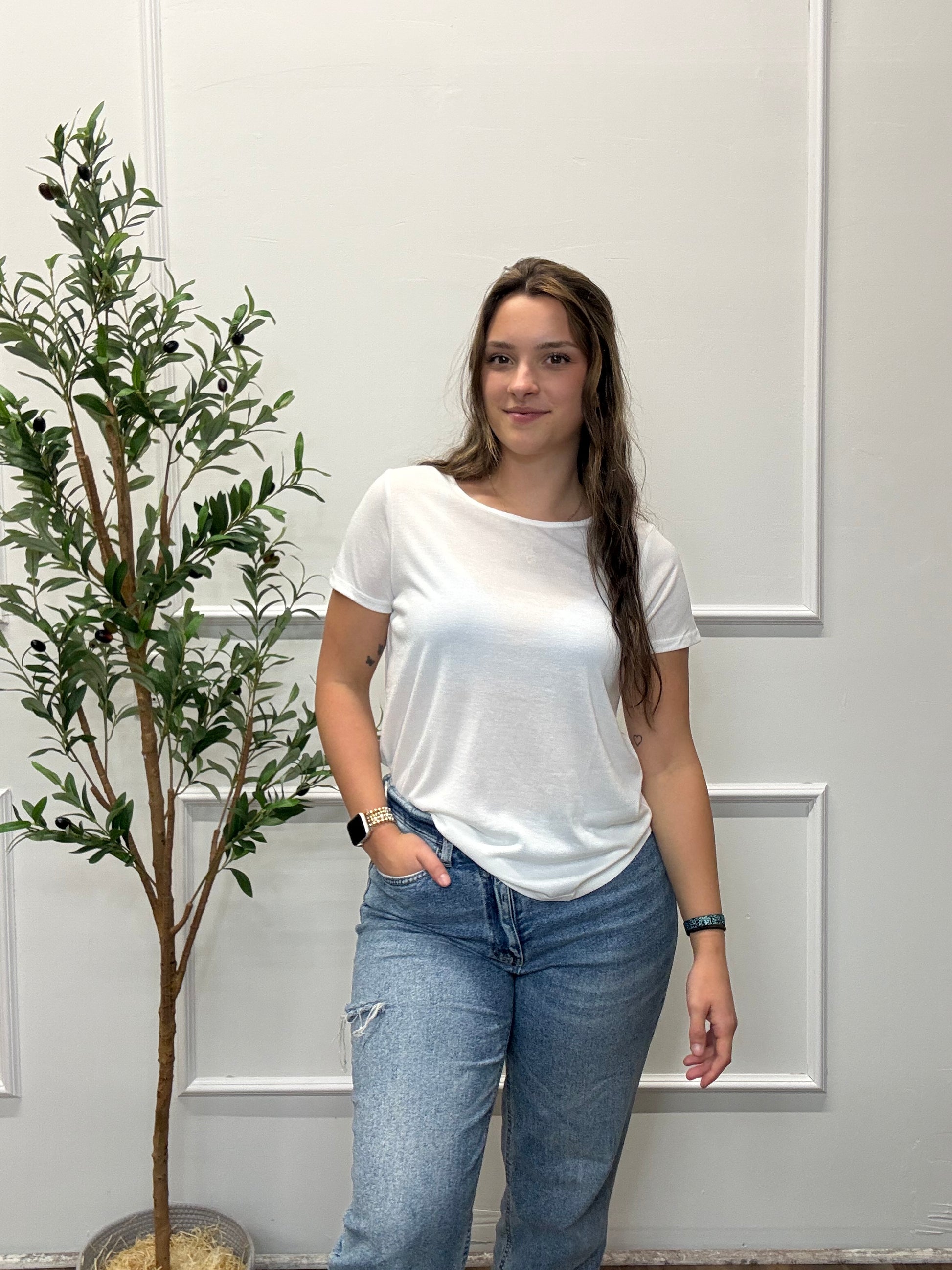 Person wearing a white t-shirt and blue jeans standing in front of a white door with a plant in the background.