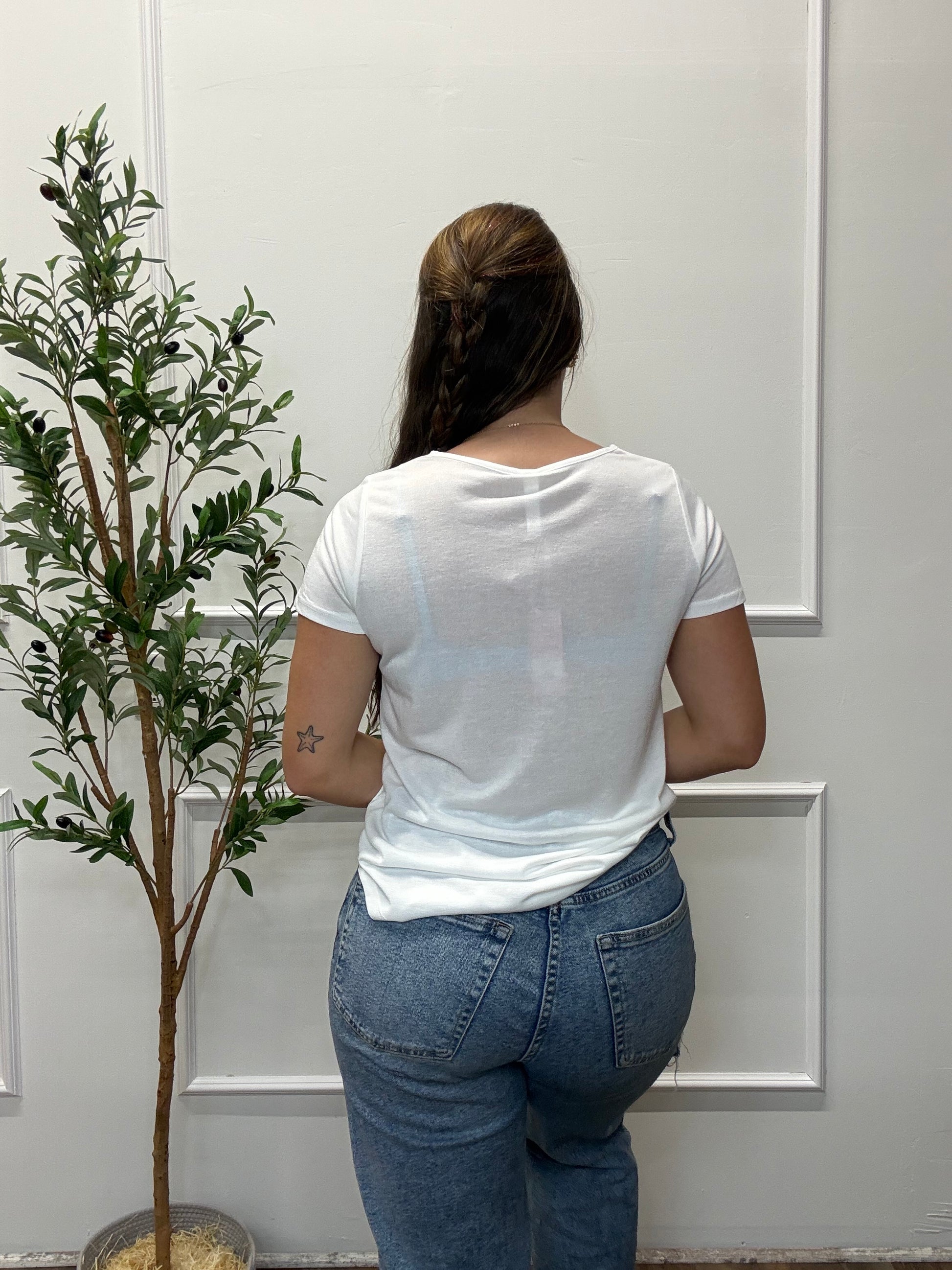 Person in white shirt and blue jeans standing against a white door with a plant.
