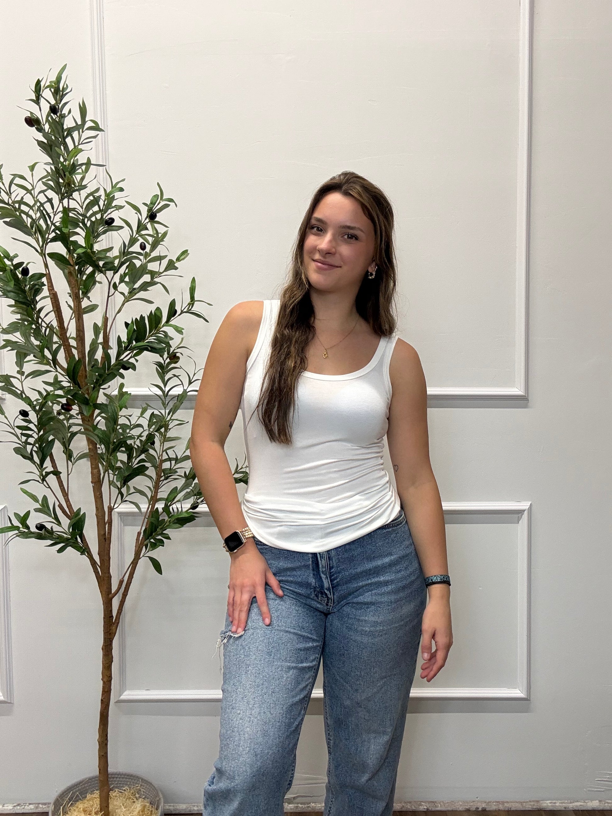 Person wearing a white top and blue jeans standing in front of a white door with a plant in the background.