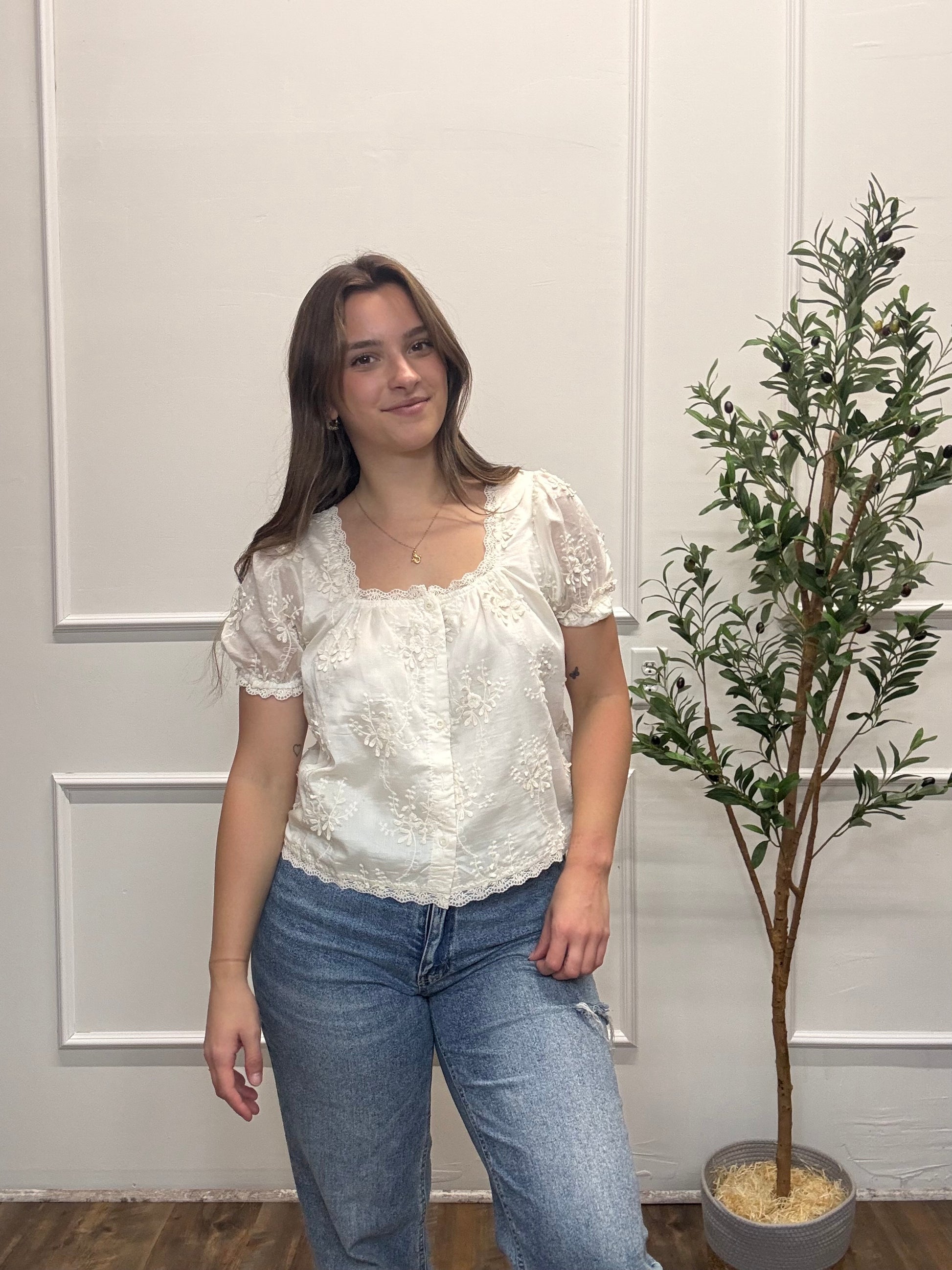 Woman wearing a white lace top and blue jeans standing in front of a white wall with a plant.