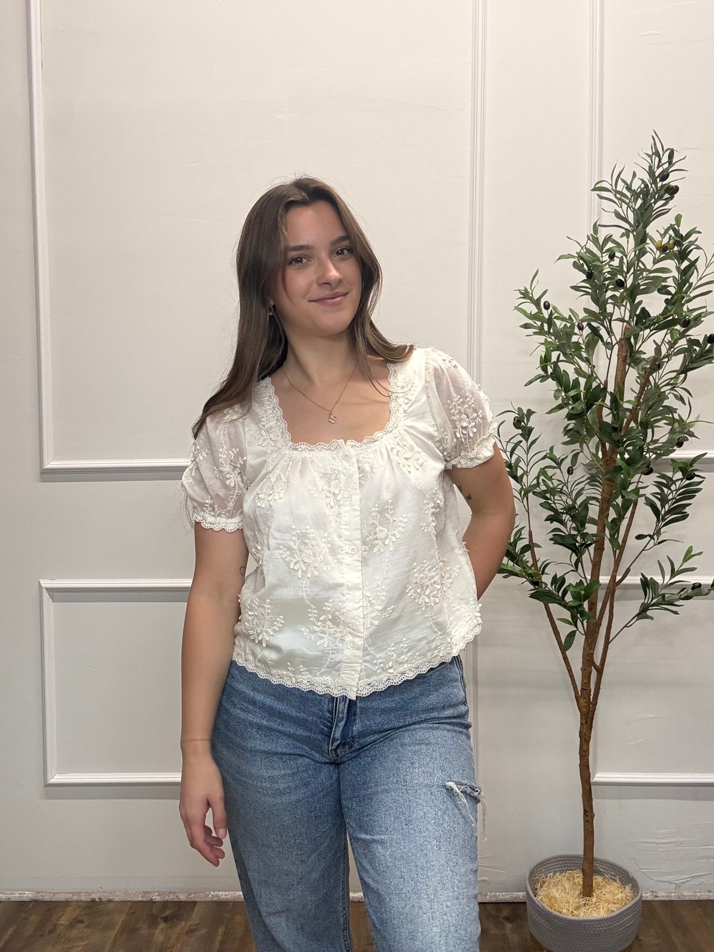 Woman wearing a white blouse and blue jeans standing next to a potted plant against a neutral wall.