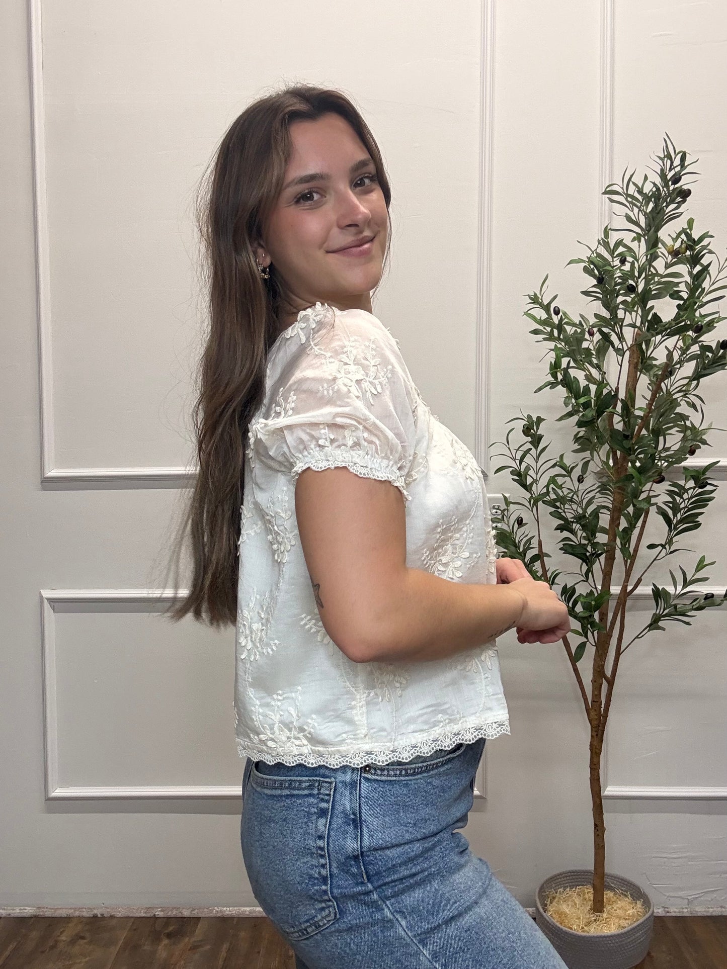 Woman wearing a white lace top and blue jeans standing next to a plant indoors.