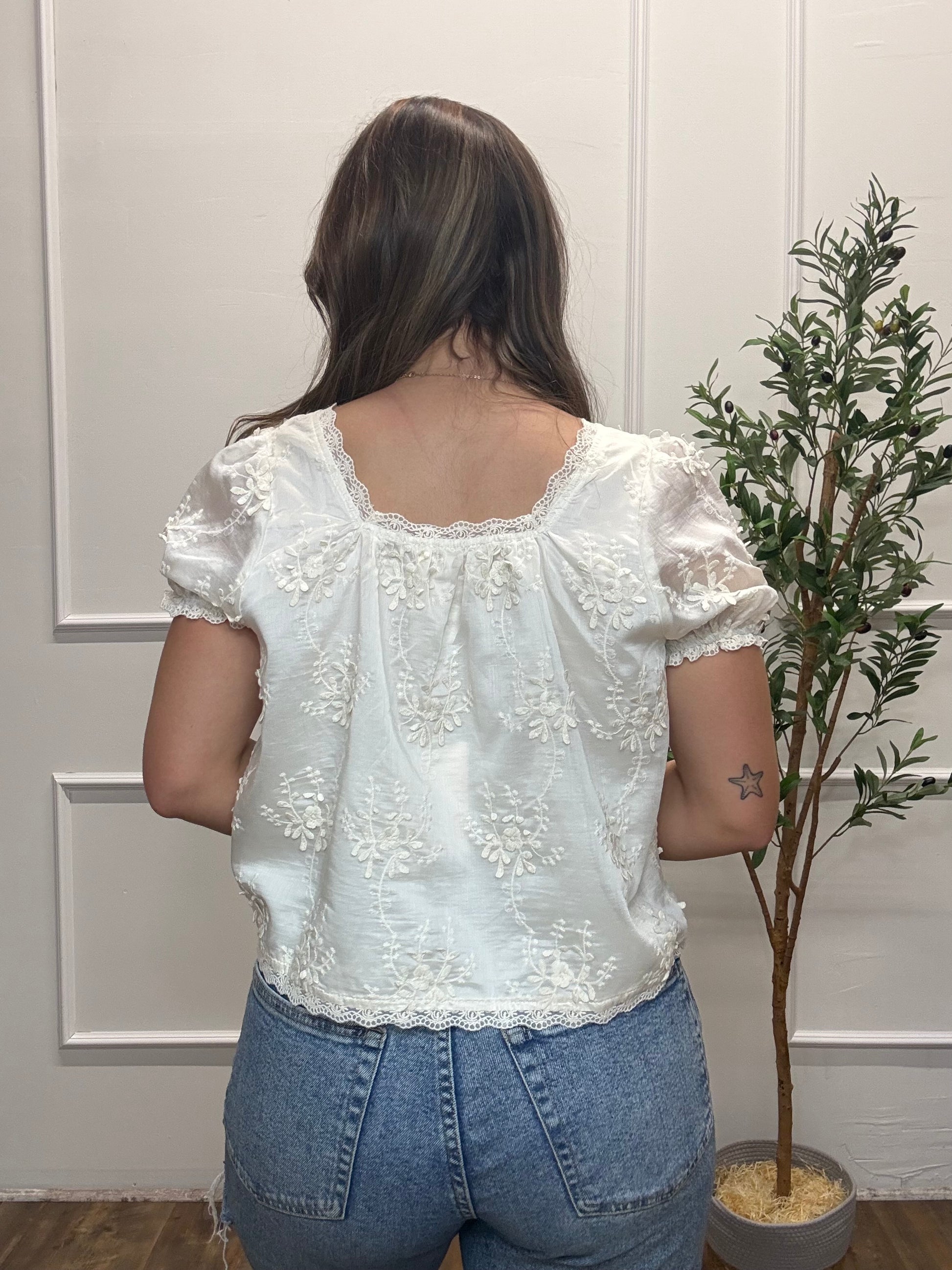 Person wearing a white lace blouse and blue jeans standing in front of a neutral wall with a plant.