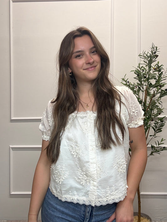 Woman wearing a white lace top and blue jeans standing indoors with a plant in the background