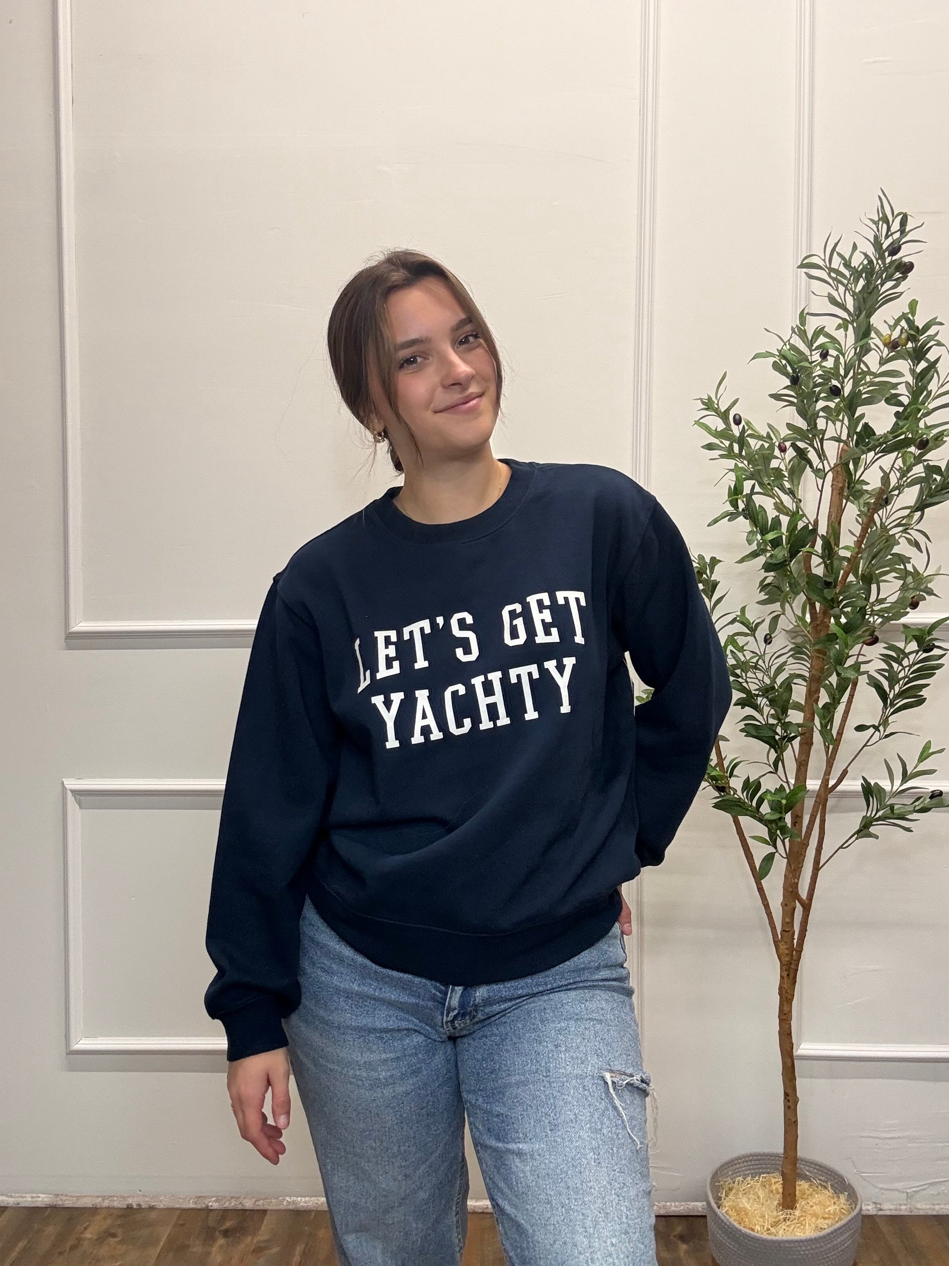 Person wearing a navy sweatshirt with 'LET'S GET YACHTY' text, standing indoors next to a plant.