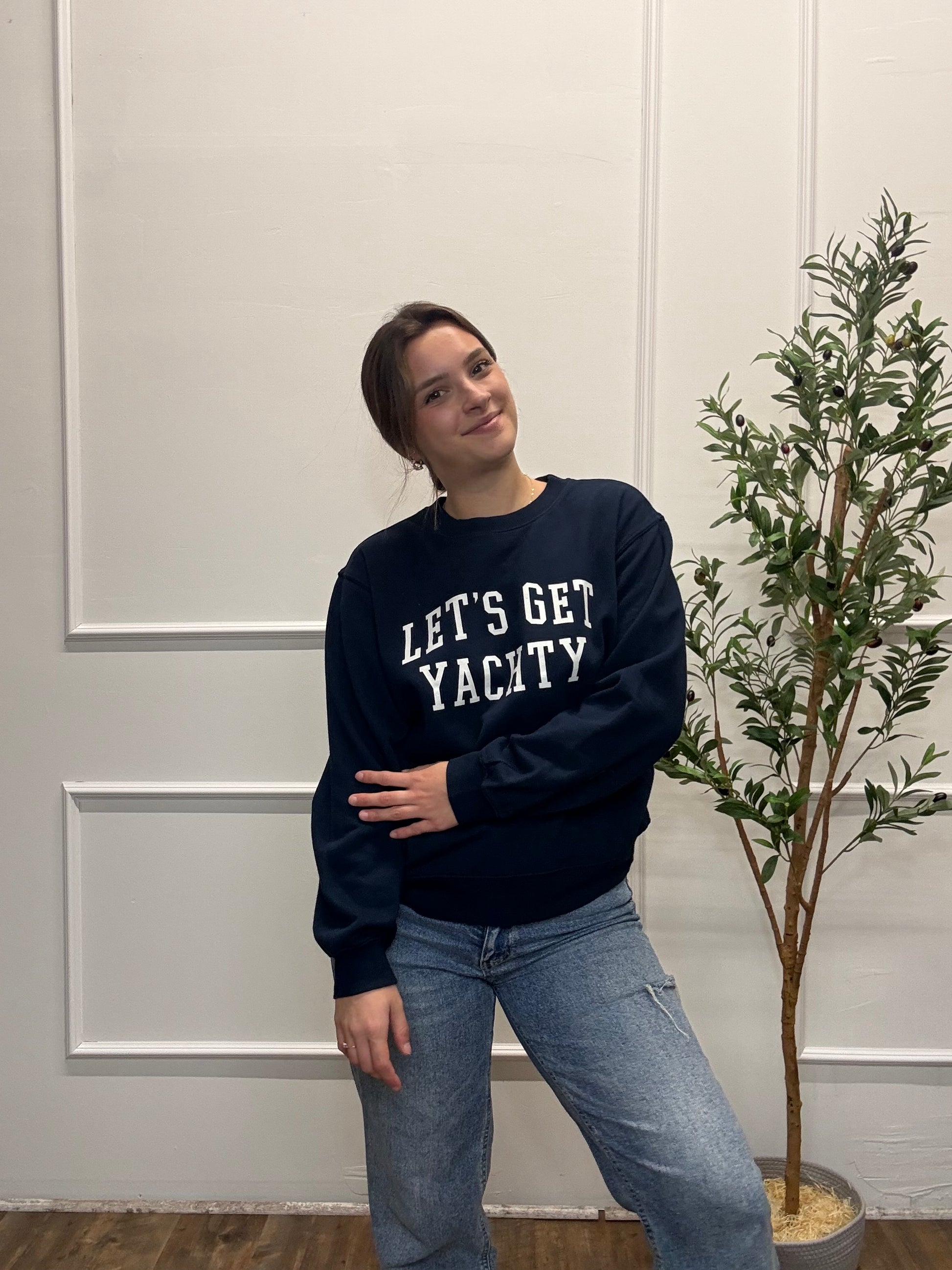 Person wearing a navy sweatshirt with 'LET'S GET YACTY' text, standing in front of a white paneled wall with a plant.