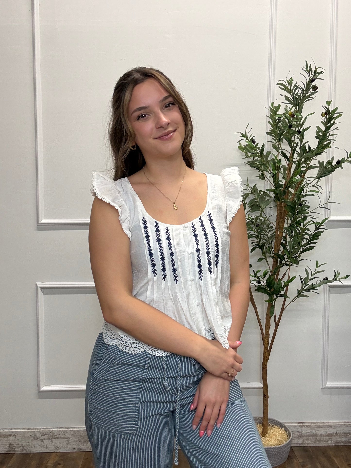 Woman wearing a white blouse with black embroidery and blue pants standing in a room with a plant.