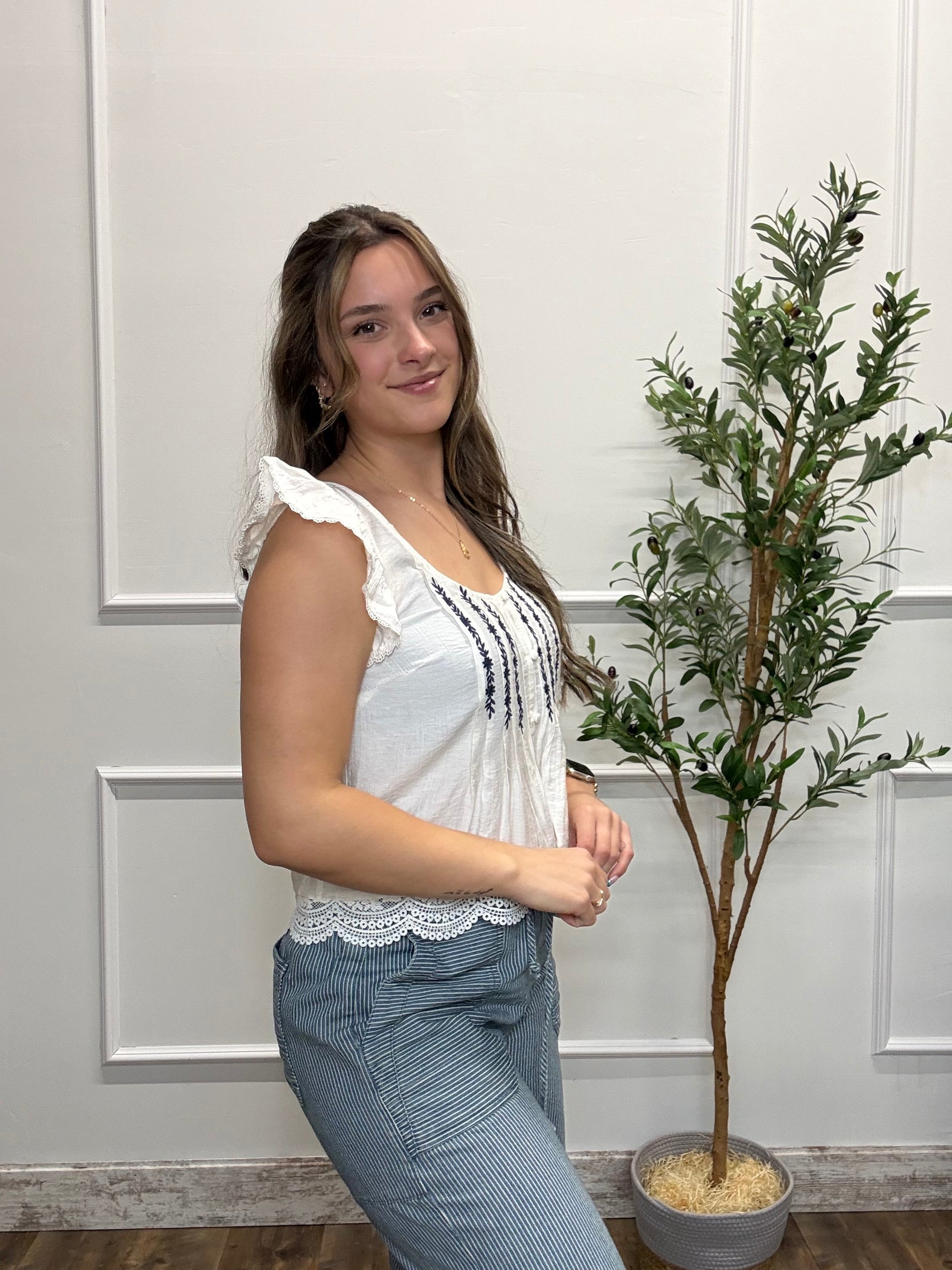 Woman in a white blouse and blue pants standing next to a potted plant against a white paneled wall.