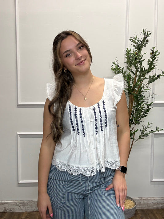 Woman wearing a white blouse with black embroidery and blue pants standing indoors.