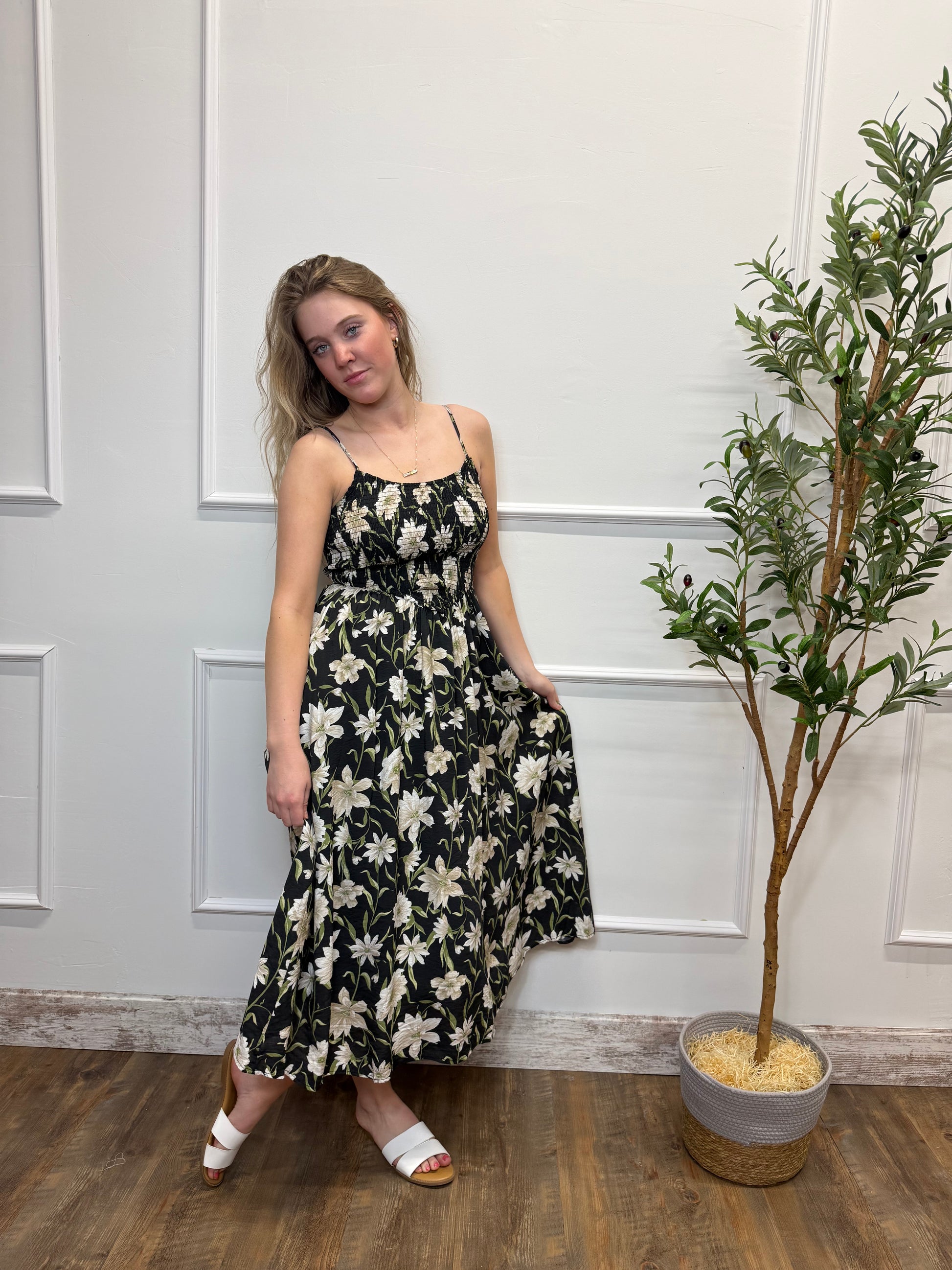 Woman in a floral dress standing next to a potted plant indoors.