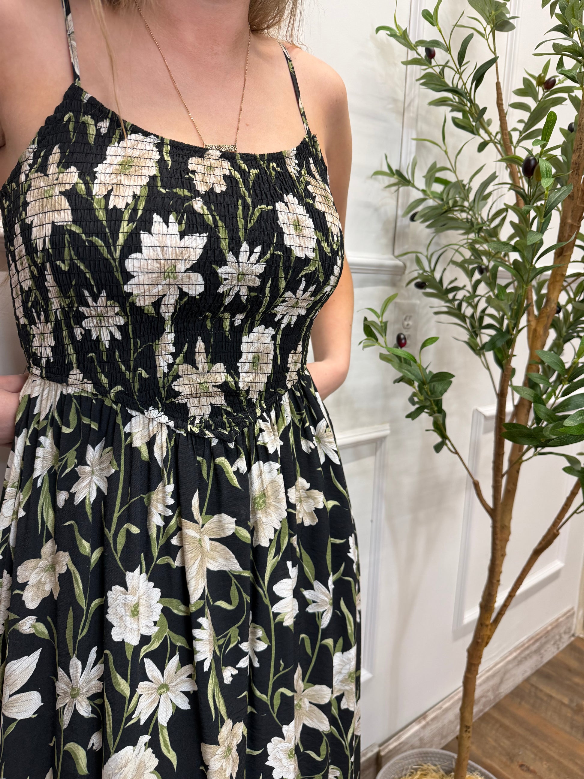 Person wearing a black floral dress with white flowers, sitting on a white surface with greenery in the background.