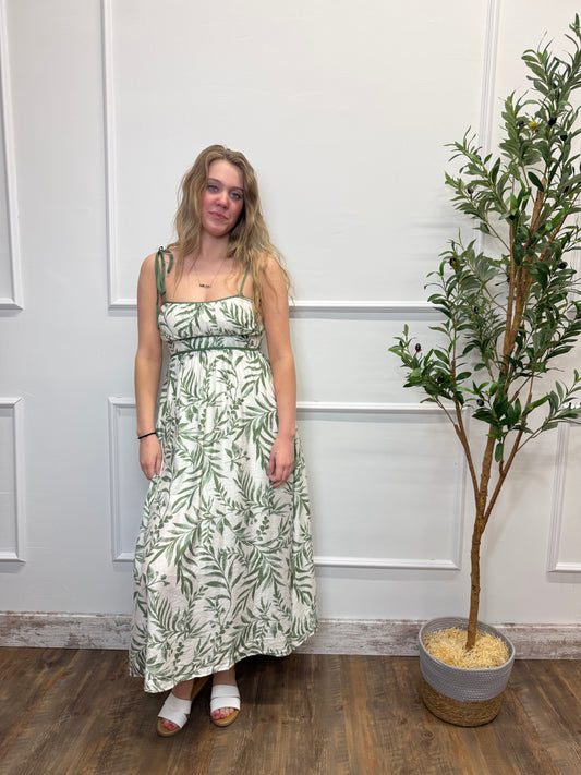 Woman in a green floral dress standing in front of a white paneled wall with a plant.