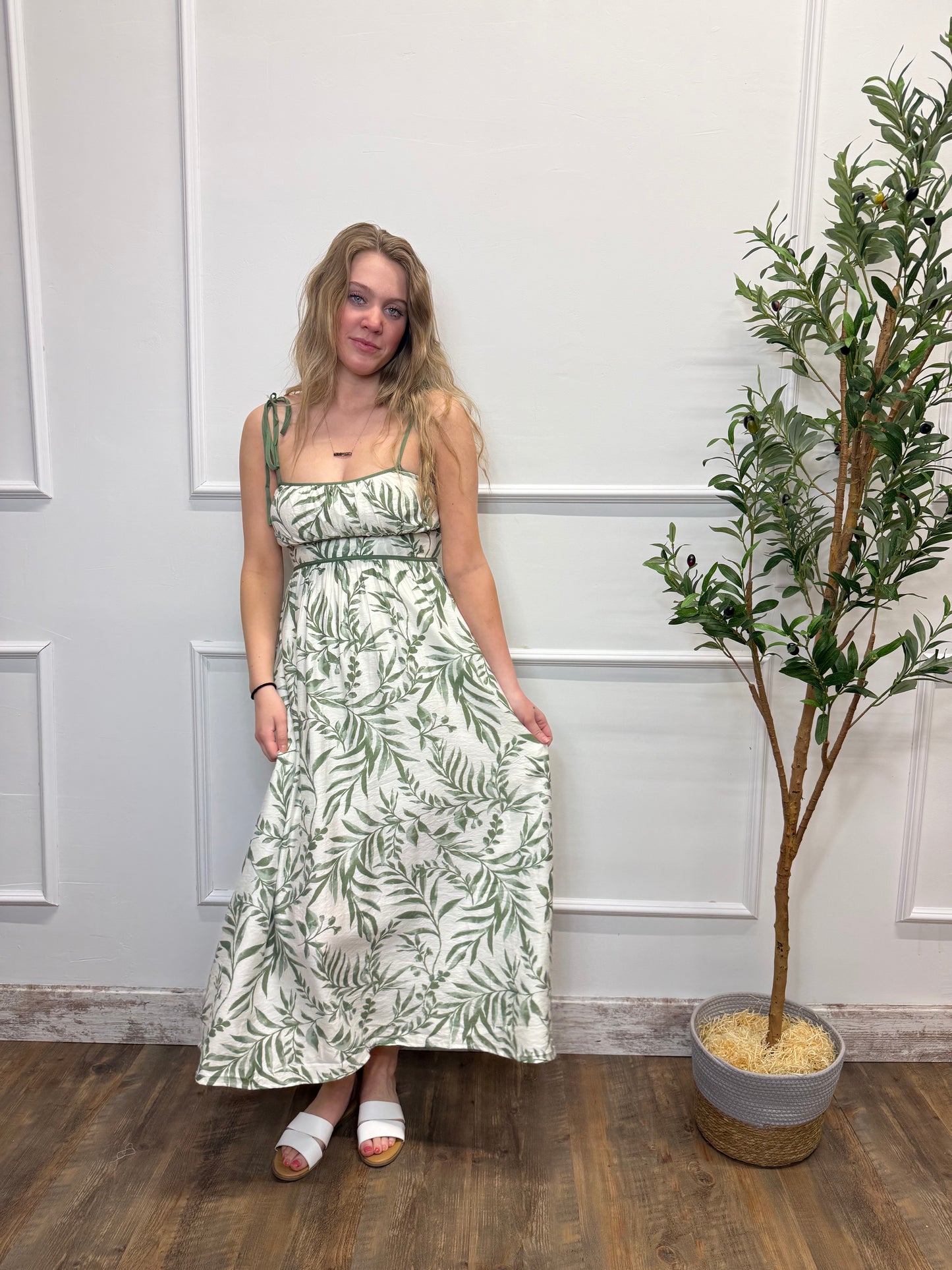 Woman in a green leaf-patterned dress standing in a room with a plant and wooden floor.