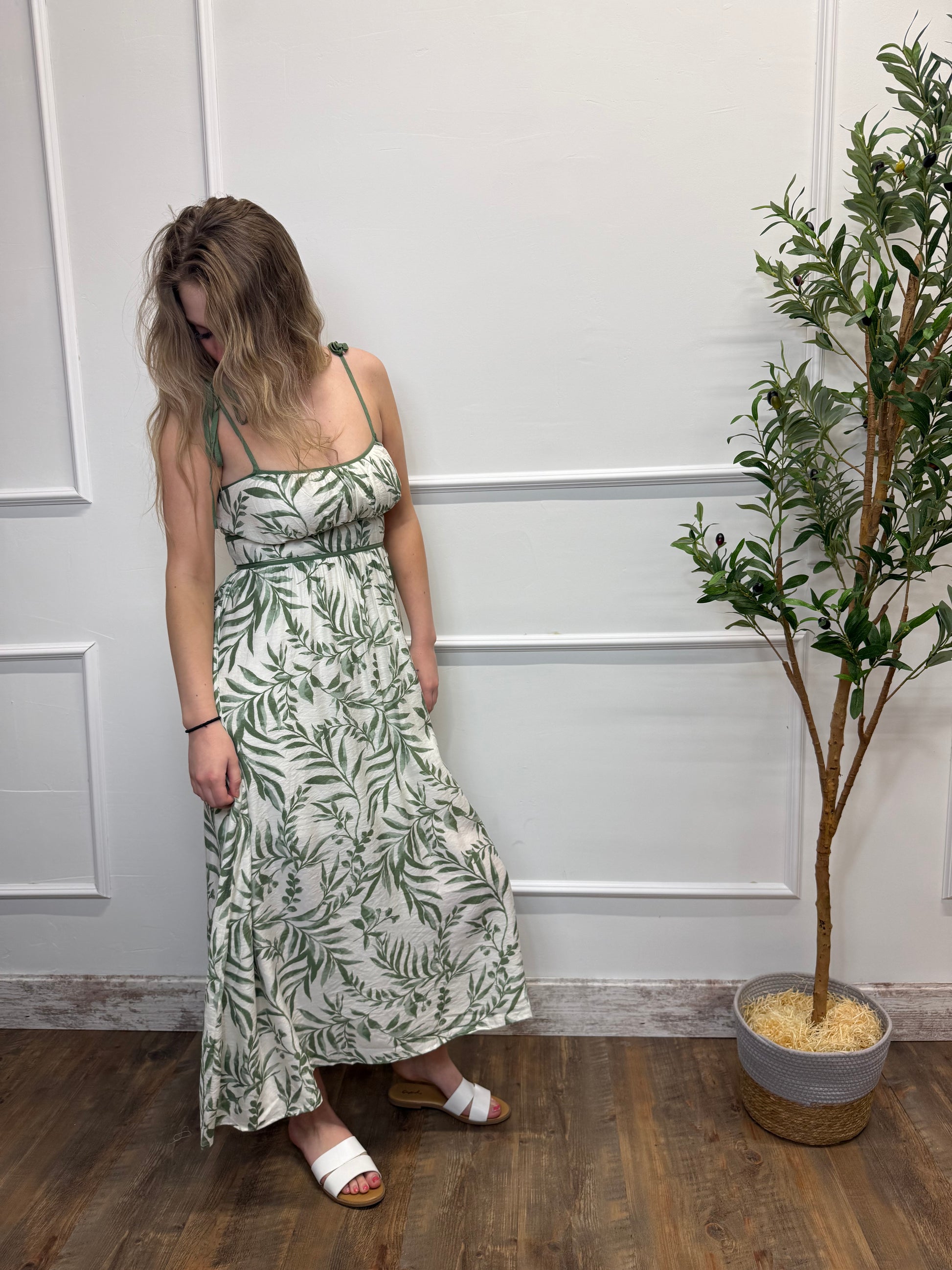 Woman in a green leaf-patterned dress standing against a white paneled wall with a plant.