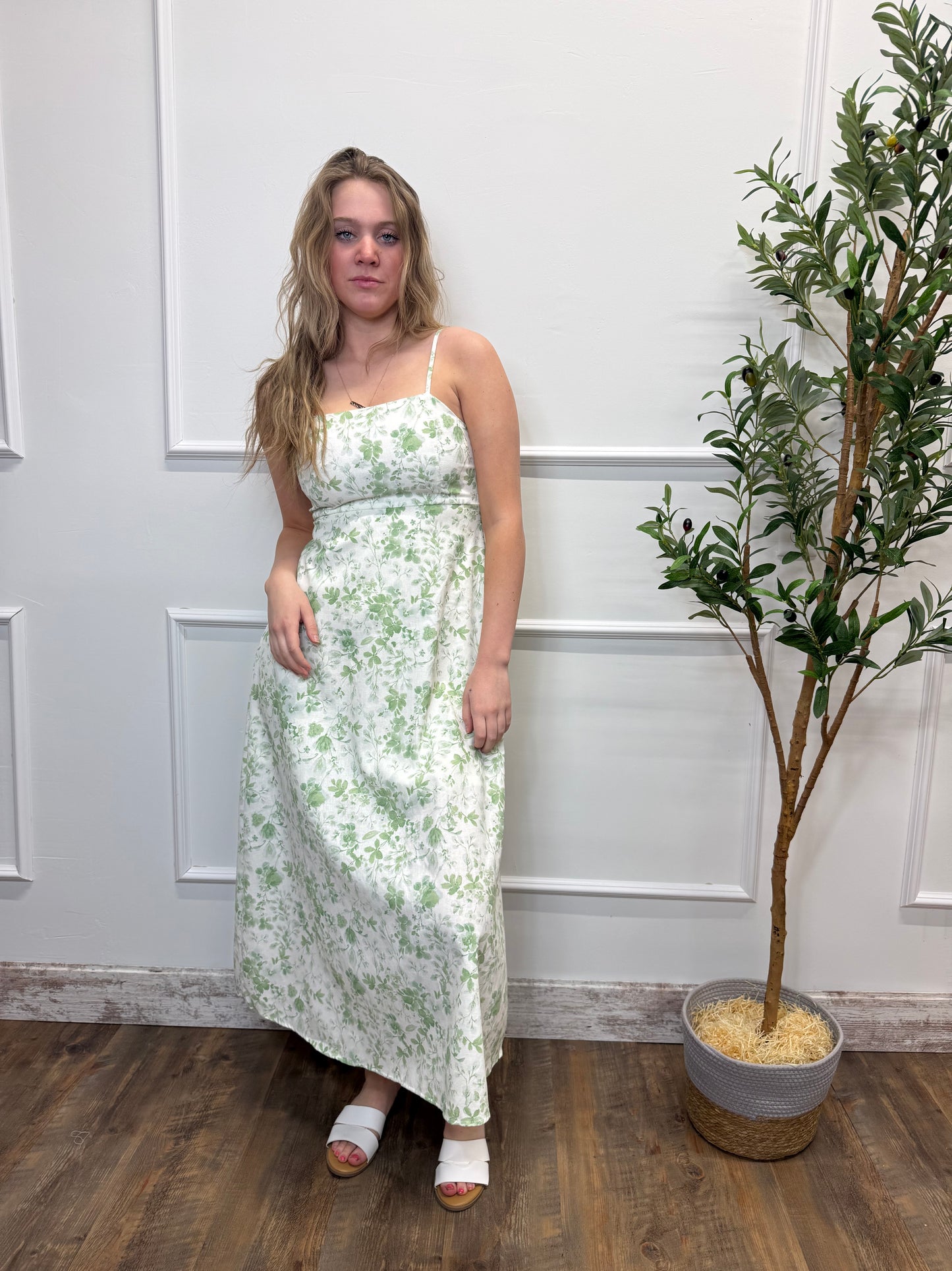 Woman in a floral dress standing in a room with a plant and wooden floor.