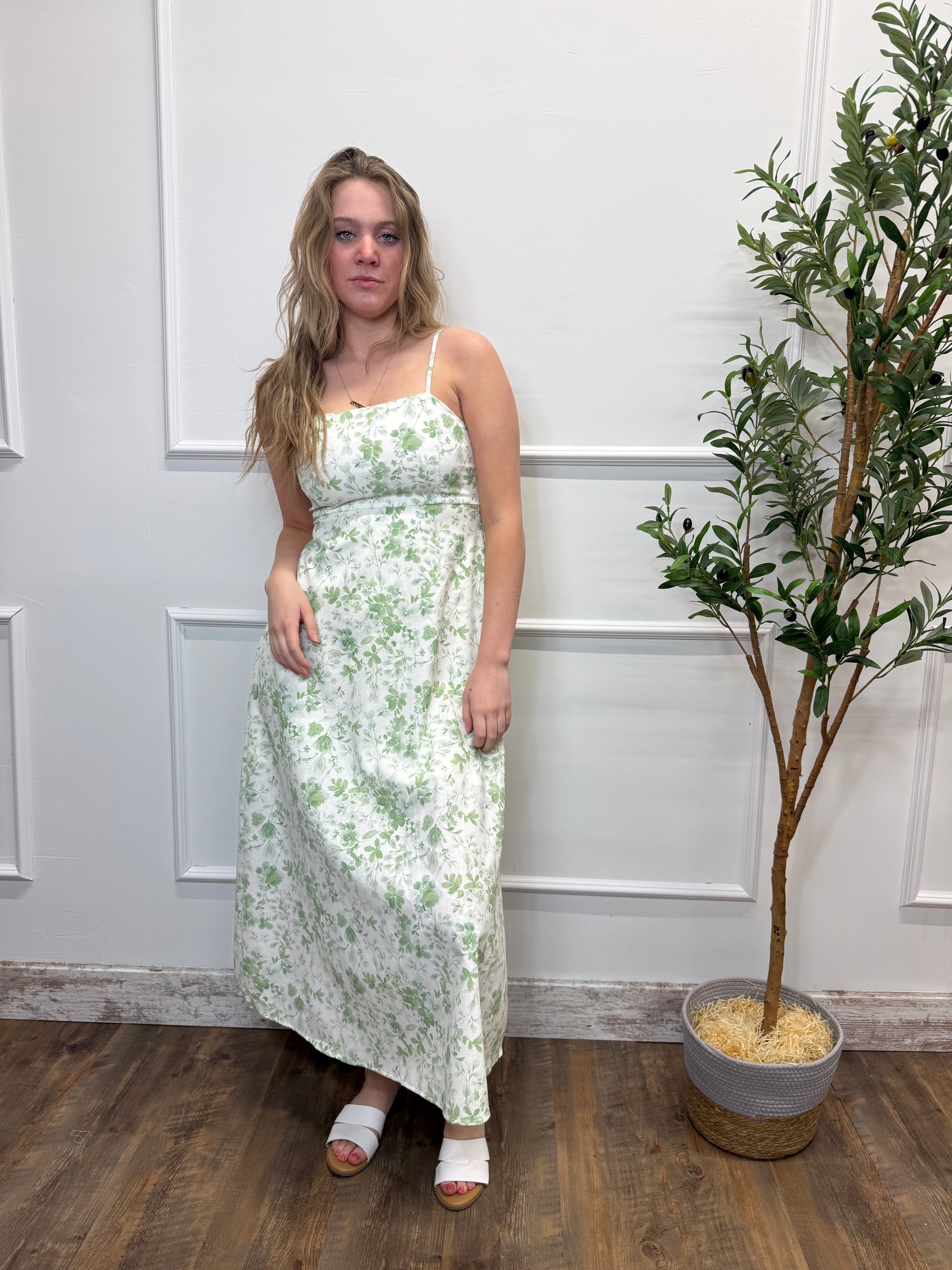 Woman in a floral dress standing in a room with a plant and wooden floor.
