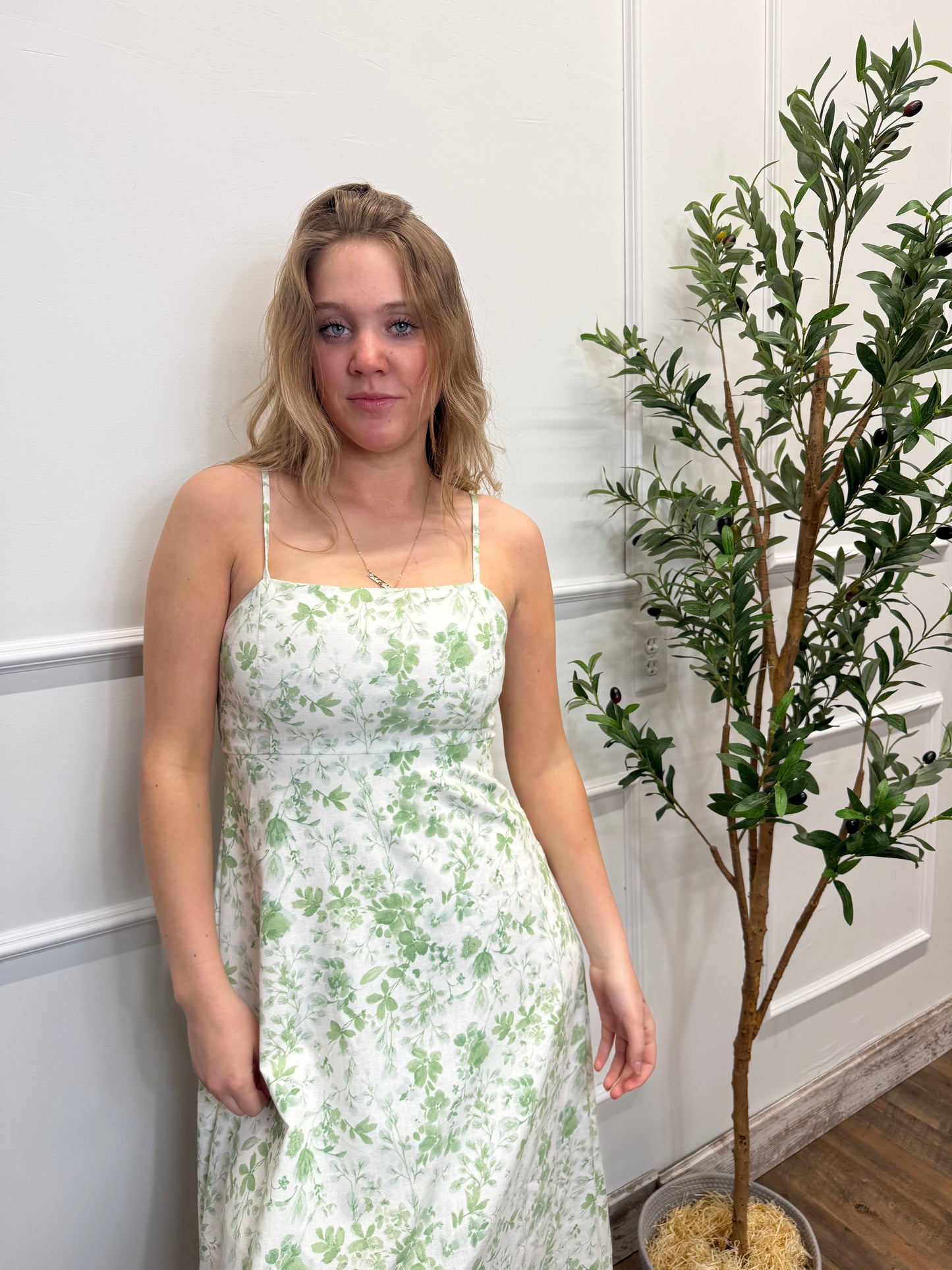 Woman in a floral dress standing next to a plant on a white wall.