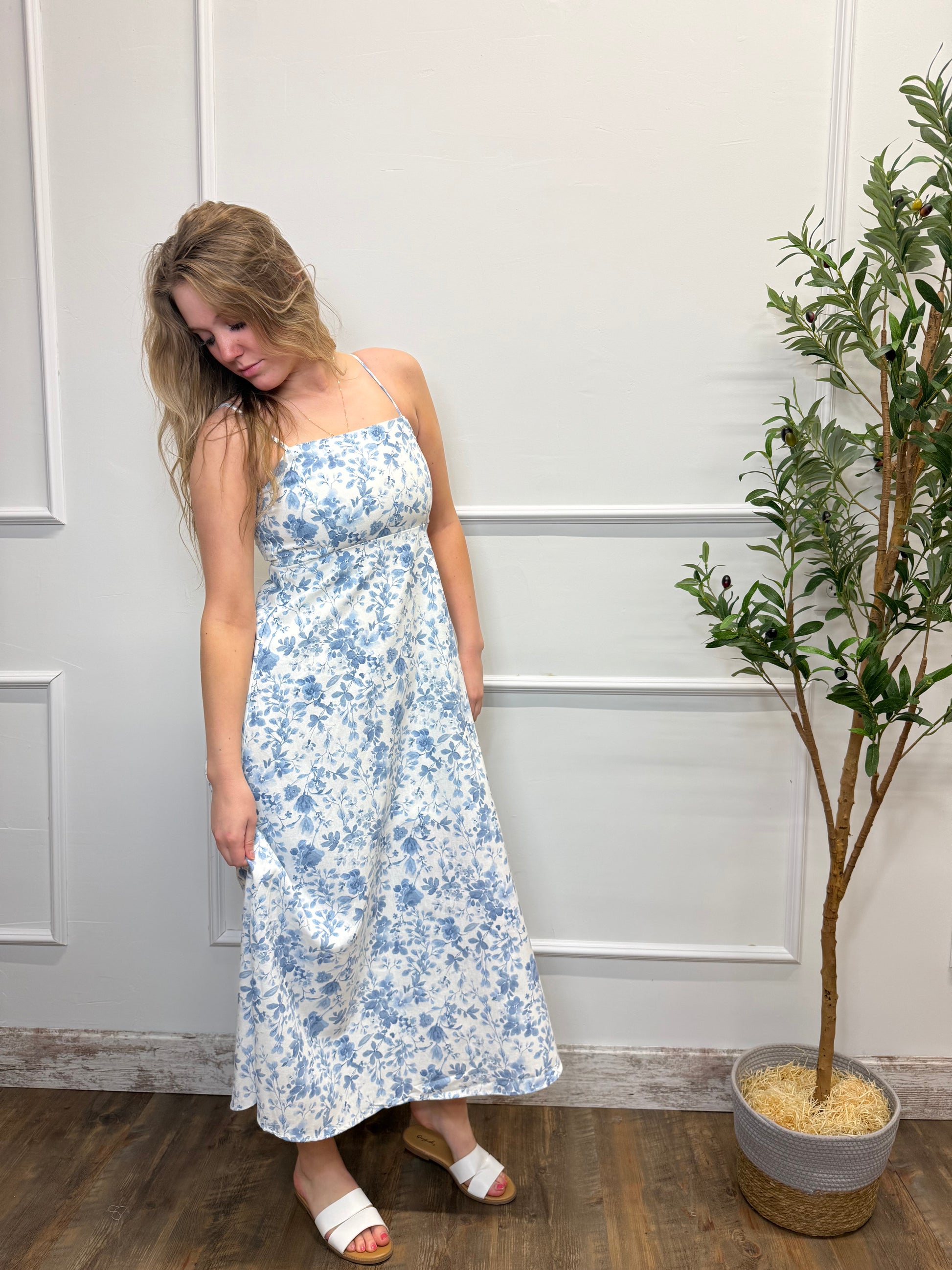 Woman wearing a blue floral dress standing in a room with a plant and wooden floor.