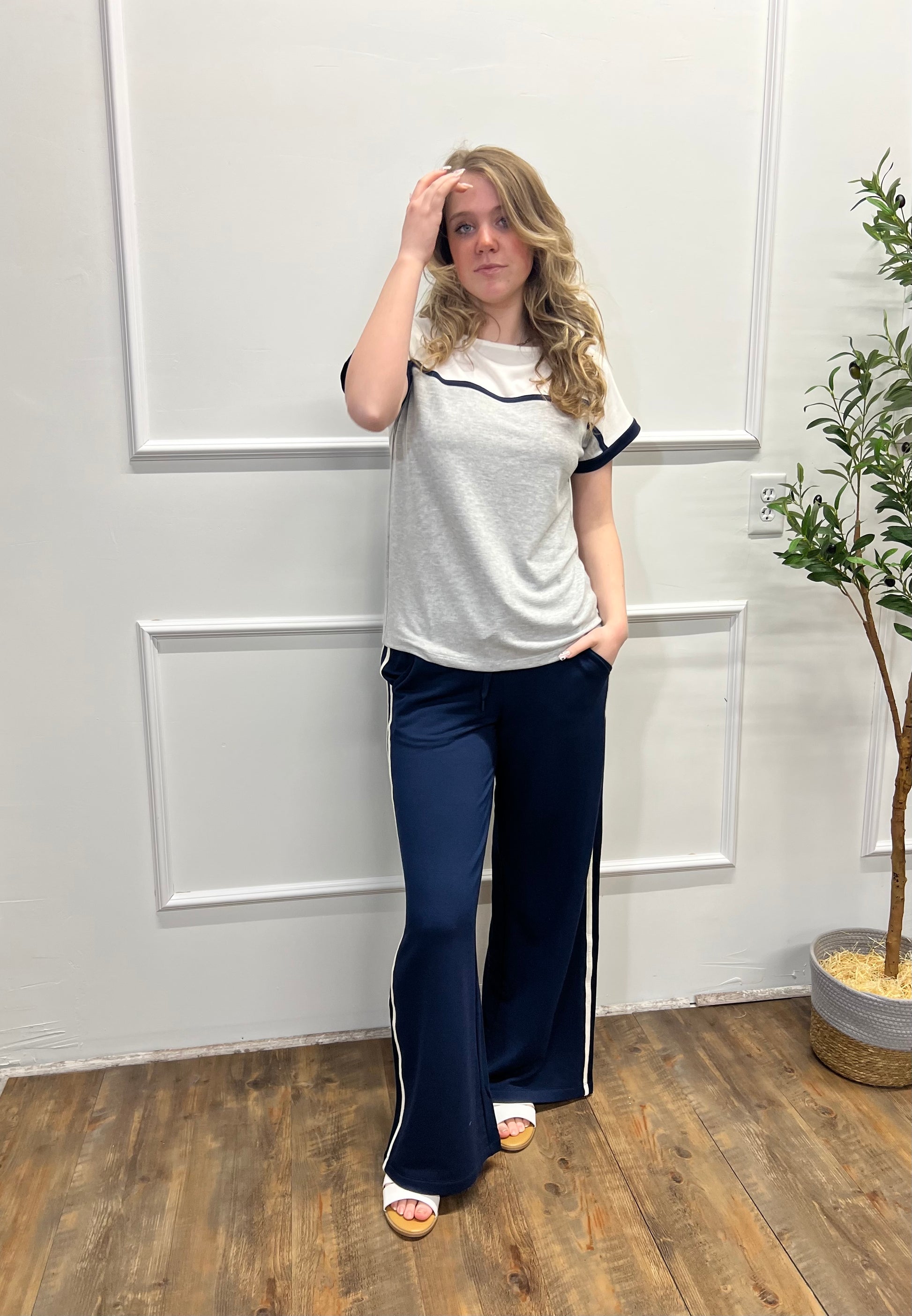 Woman wearing a white top with black trim and blue pants in a room with wooden flooring and a plant.