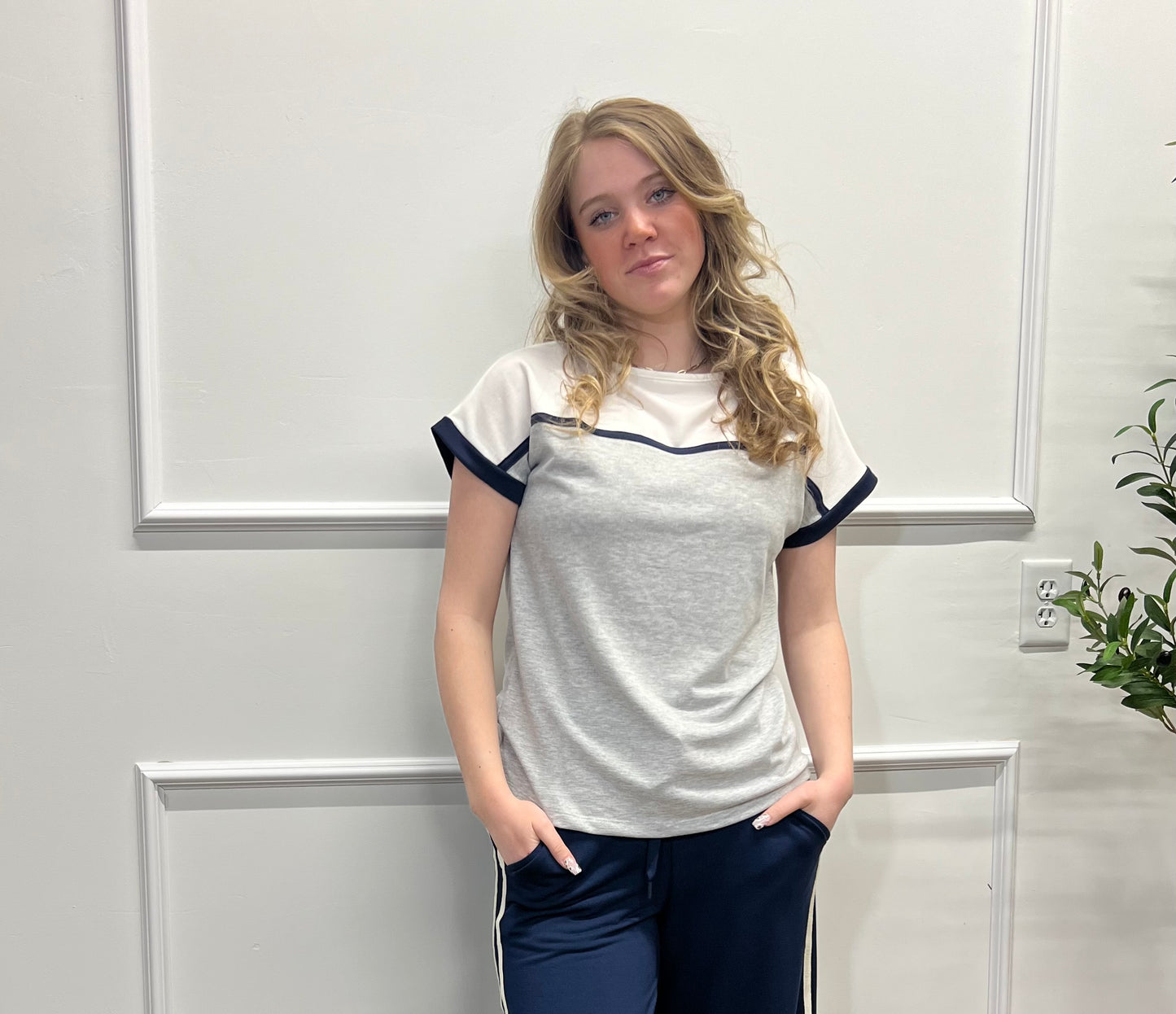 Woman wearing a white t-shirt with navy accents in a room with a white wall and plant.