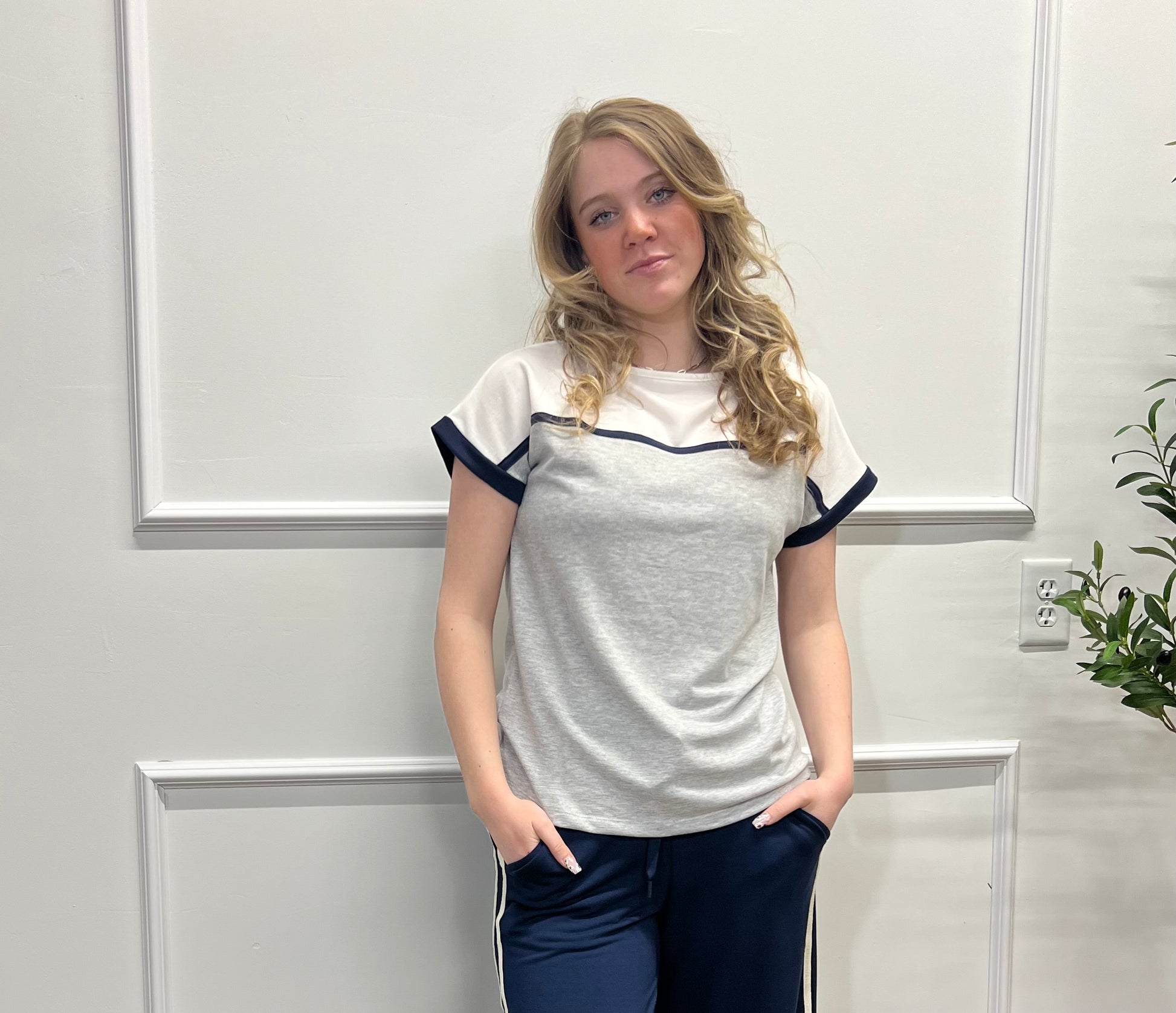 Woman wearing a white t-shirt with navy accents in a room with a white wall and plant.
