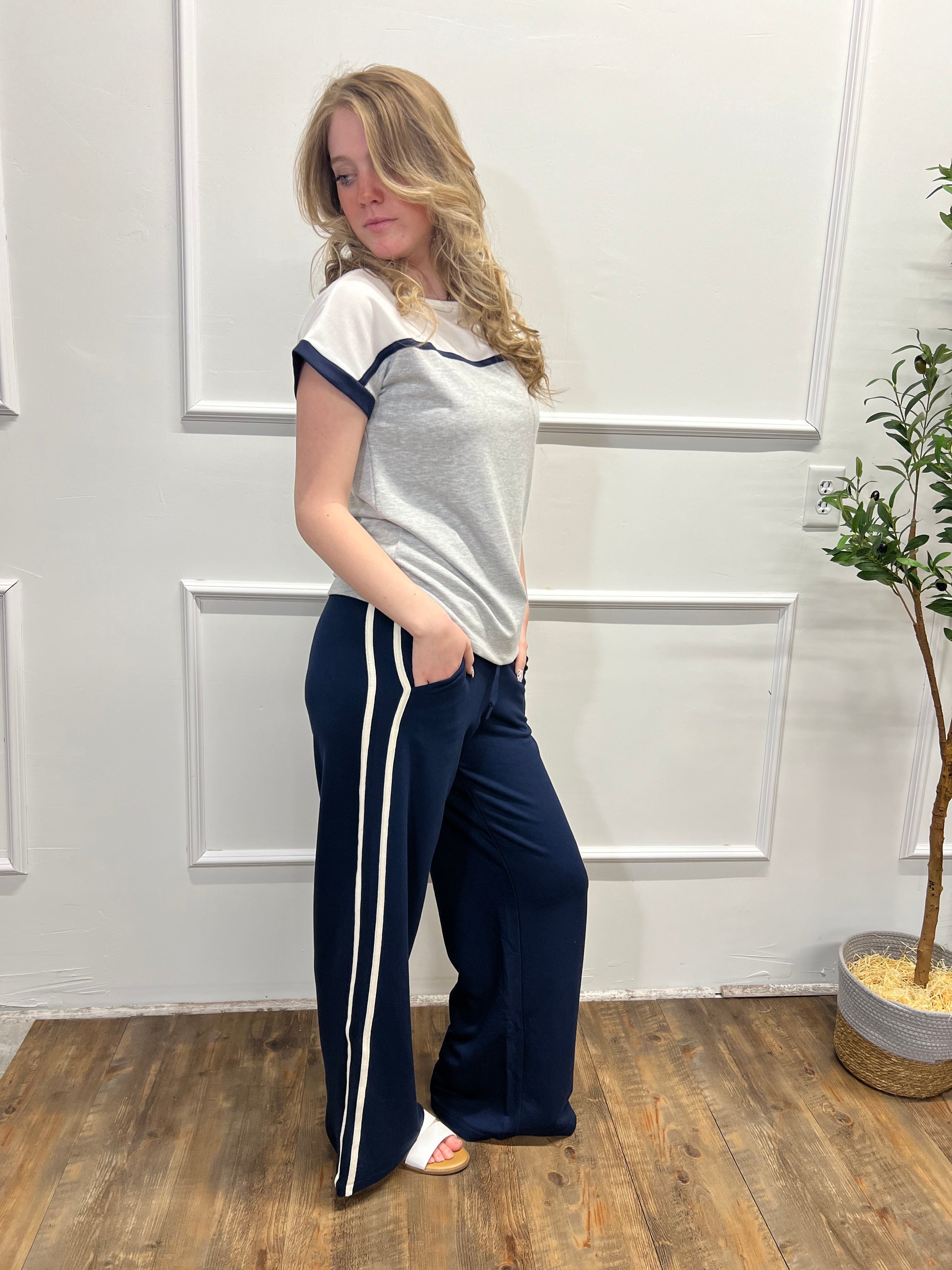 Person wearing a gray and white top with navy pants in a room with wooden flooring and a plant.