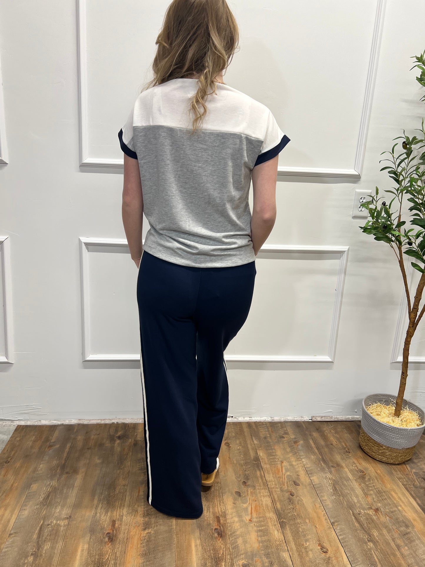Person wearing a white and gray top with navy pants in a room with wooden flooring and a plant.