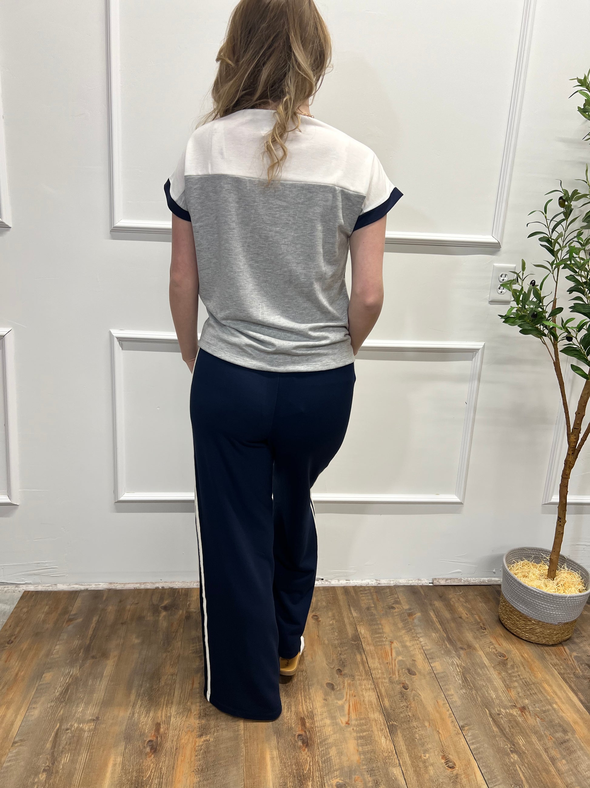 Person wearing a white and gray top with navy pants in a room with wooden flooring and a plant.