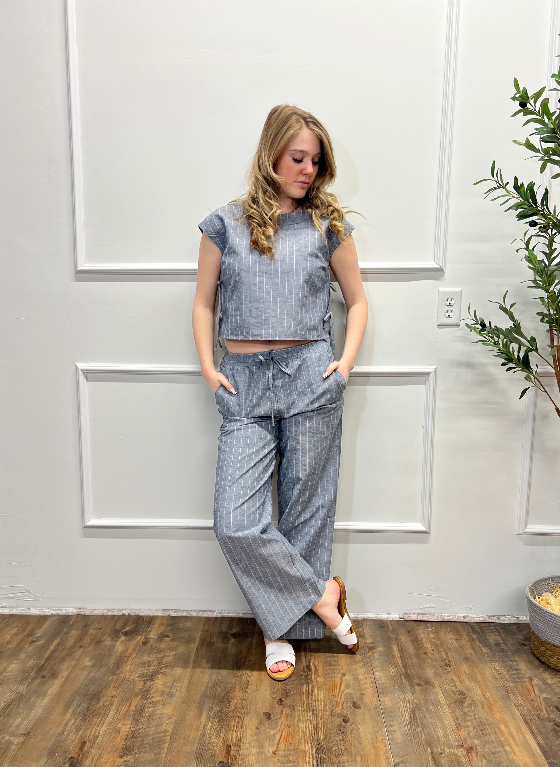 Woman wearing a gray outfit with wide-leg pants in a room with wooden flooring and a plant.