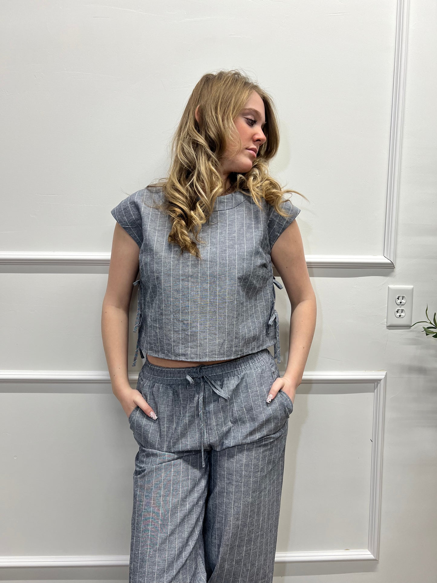 Woman wearing a sleeveless denim top and plaid pants standing against a white paneled wall.