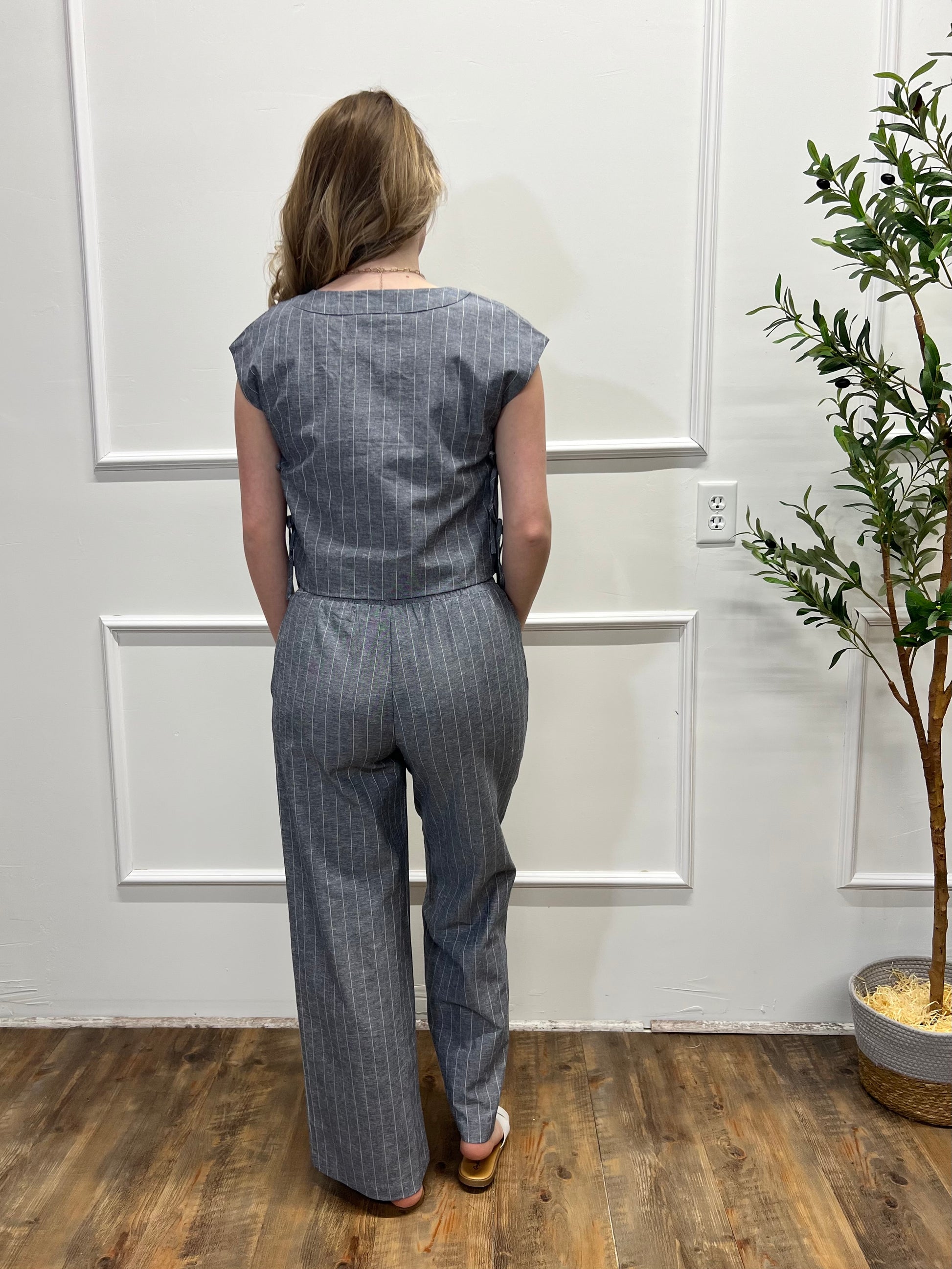 Person wearing a gray pinstripe jumpsuit standing in a room with a plant and wooden floor.
