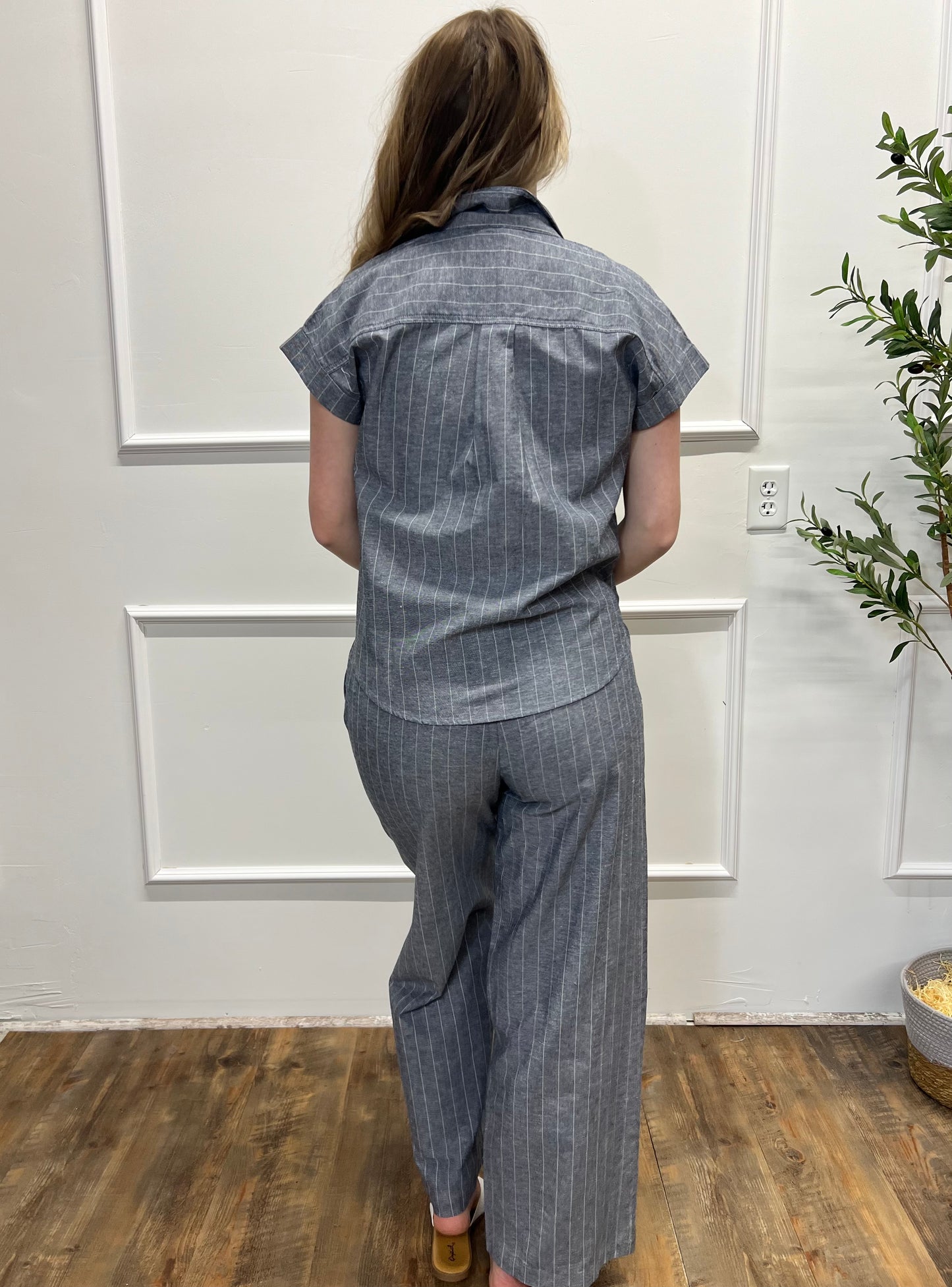 Person wearing a blue pinstripe shirt and pants in a room with wooden flooring and a plant.