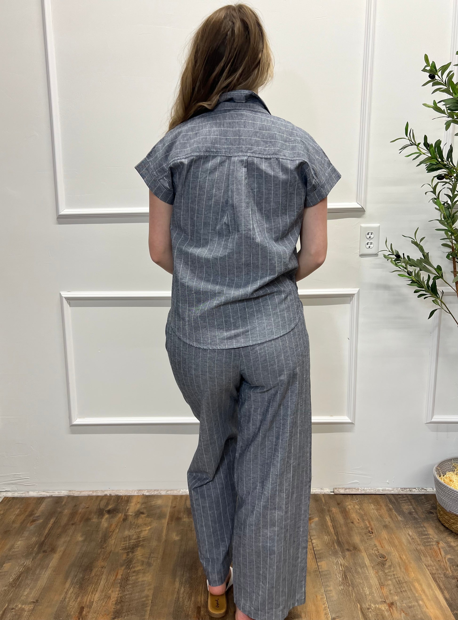 Person wearing a blue pinstripe shirt and pants in a room with wooden flooring and a plant.
