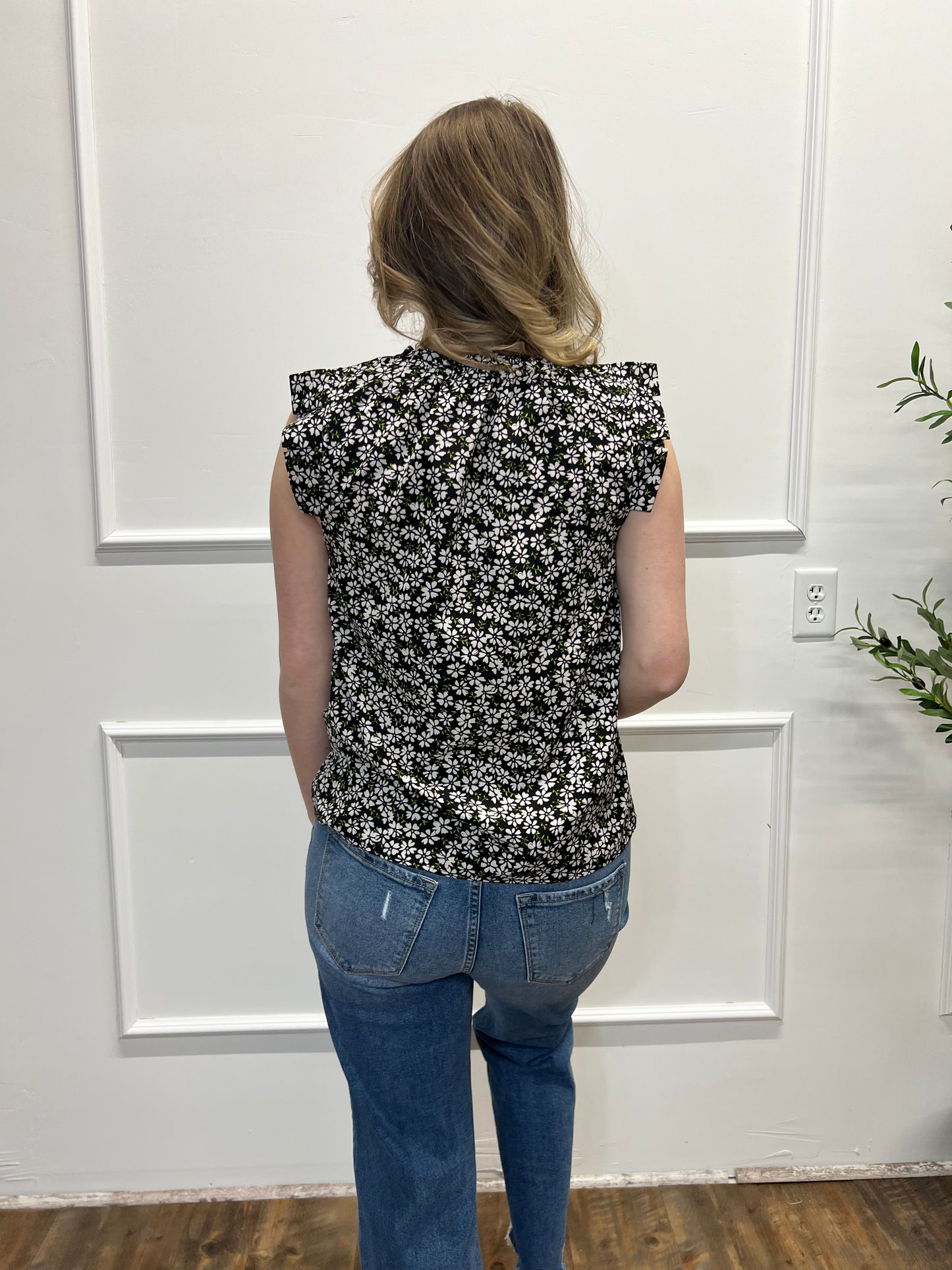 Person wearing a black floral top and blue jeans standing in a room with white walls and a wooden floor.