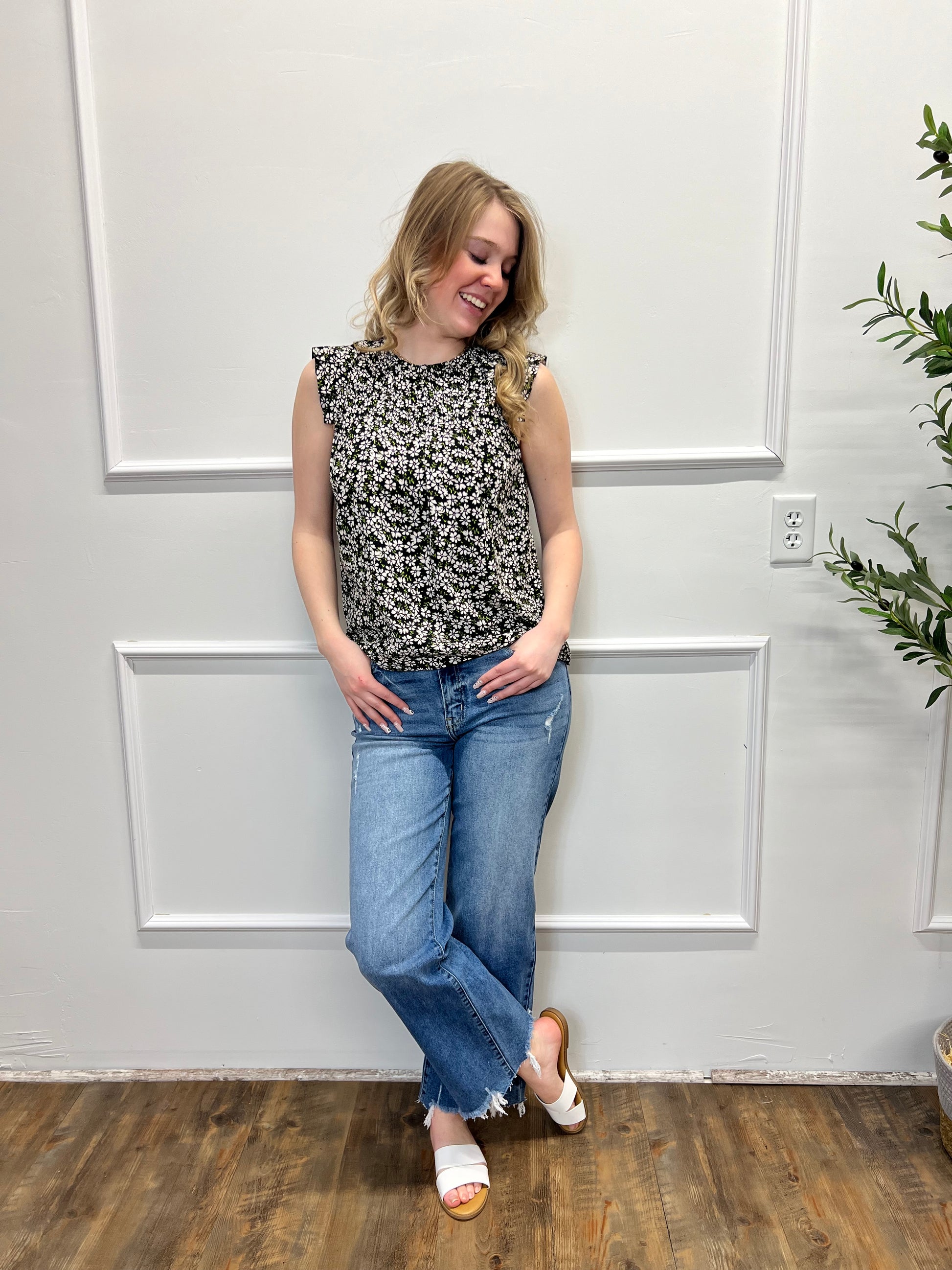 Person wearing a floral top and blue jeans standing in front of a white paneled wall.