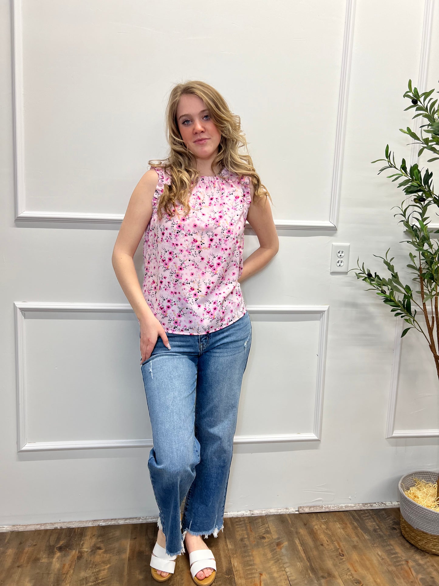 Woman in a floral top and jeans standing in front of a white door with a plant on the floor.