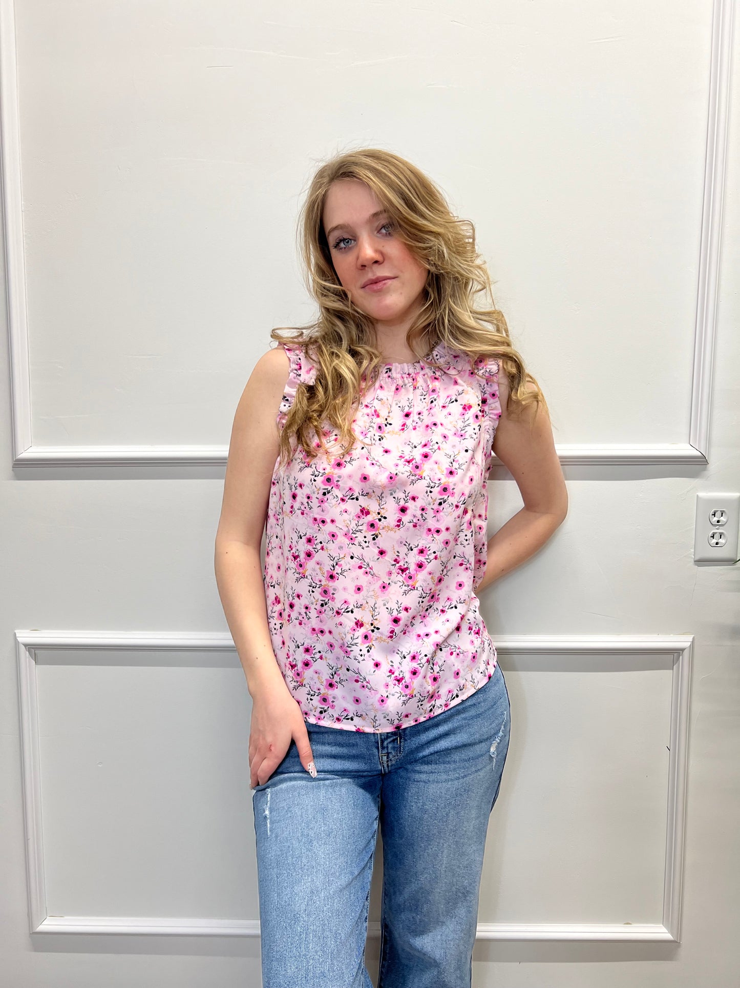 Woman wearing a pink floral top and blue jeans standing against a white paneled wall.