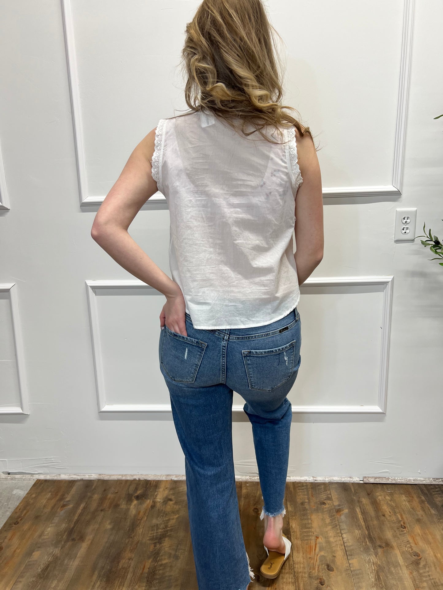 Person wearing a white sleeveless top and blue jeans standing in a room with white walls and a wooden floor.
