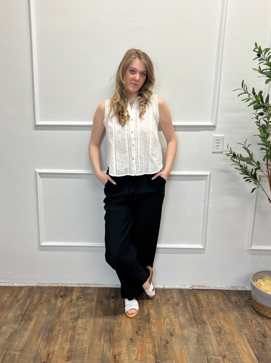 Woman wearing a white sleeveless blouse and black pants standing against a gray wall.
