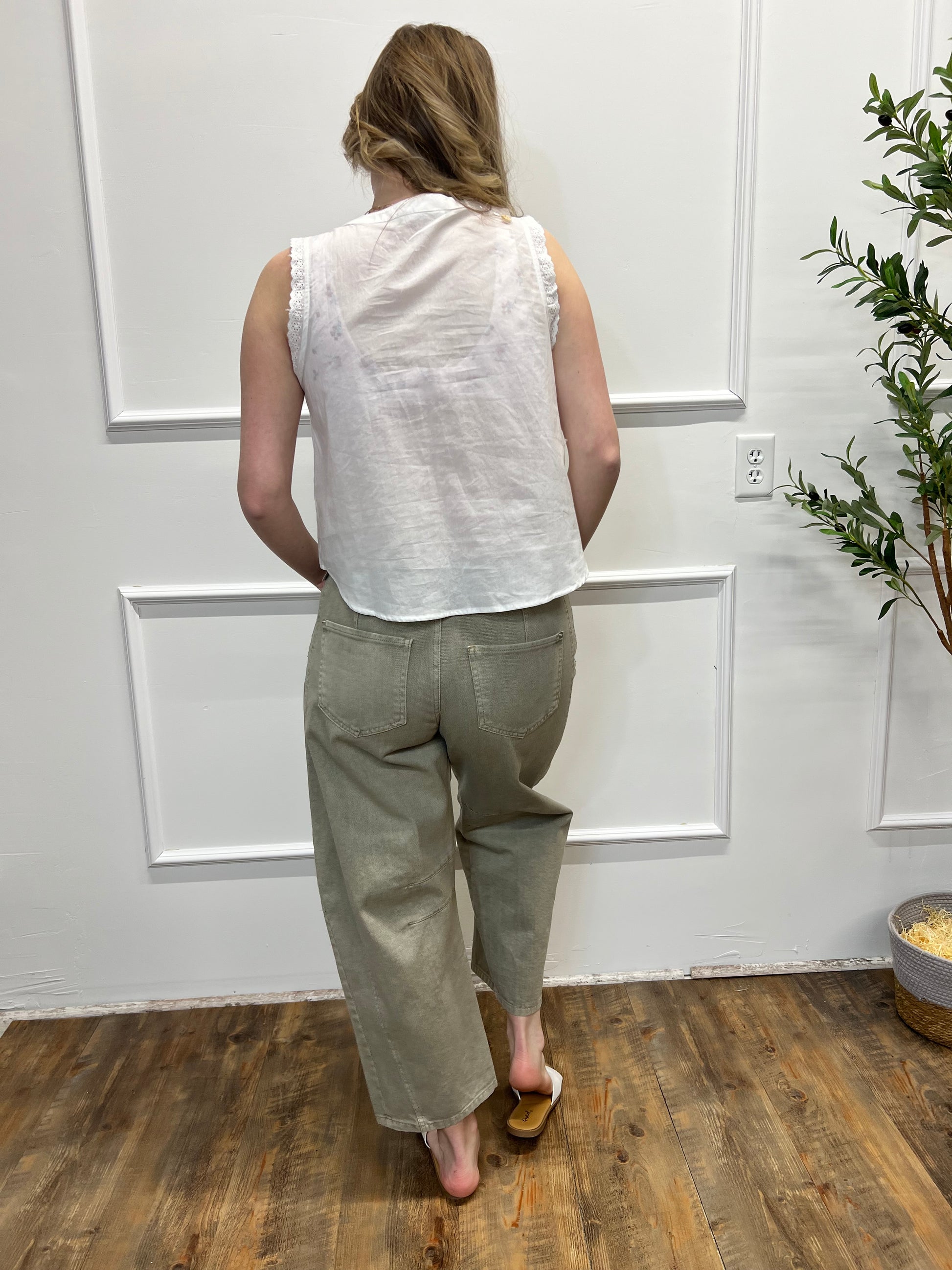 Person in a white shirt and green pants standing on a wooden floor with a white wall and plant in the background.