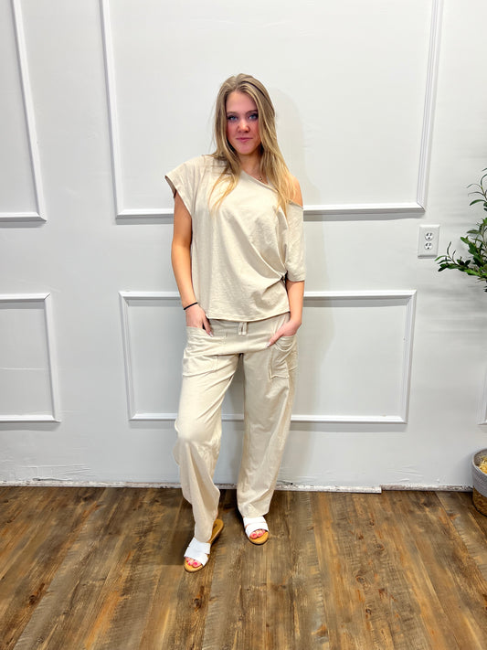 Person wearing a beige outfit standing in a room with white walls and wooden flooring.