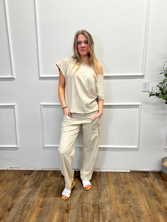 Person wearing a beige outfit standing in a room with white walls and wooden flooring.