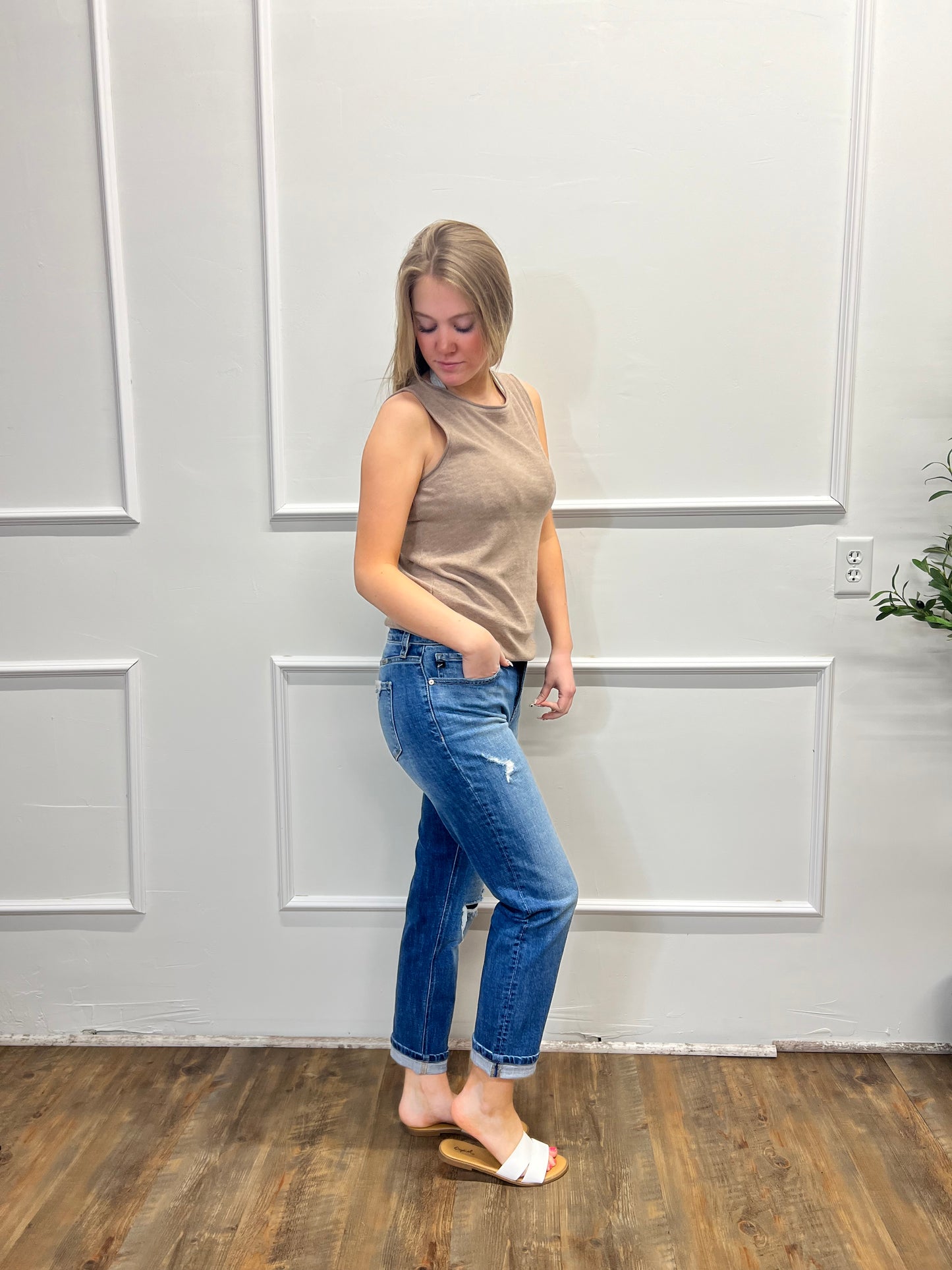 Person wearing a beige top and blue jeans standing against a white paneled wall.