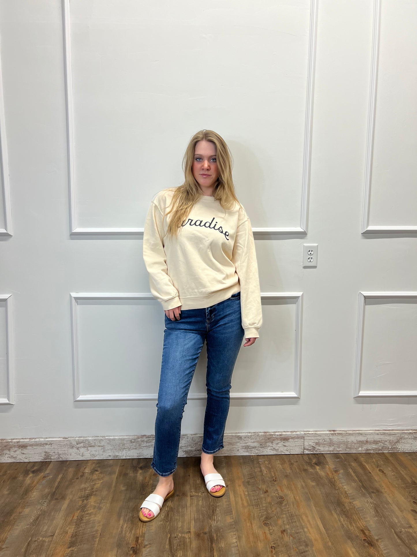 Person wearing a beige sweatshirt and blue jeans standing in front of a white door.